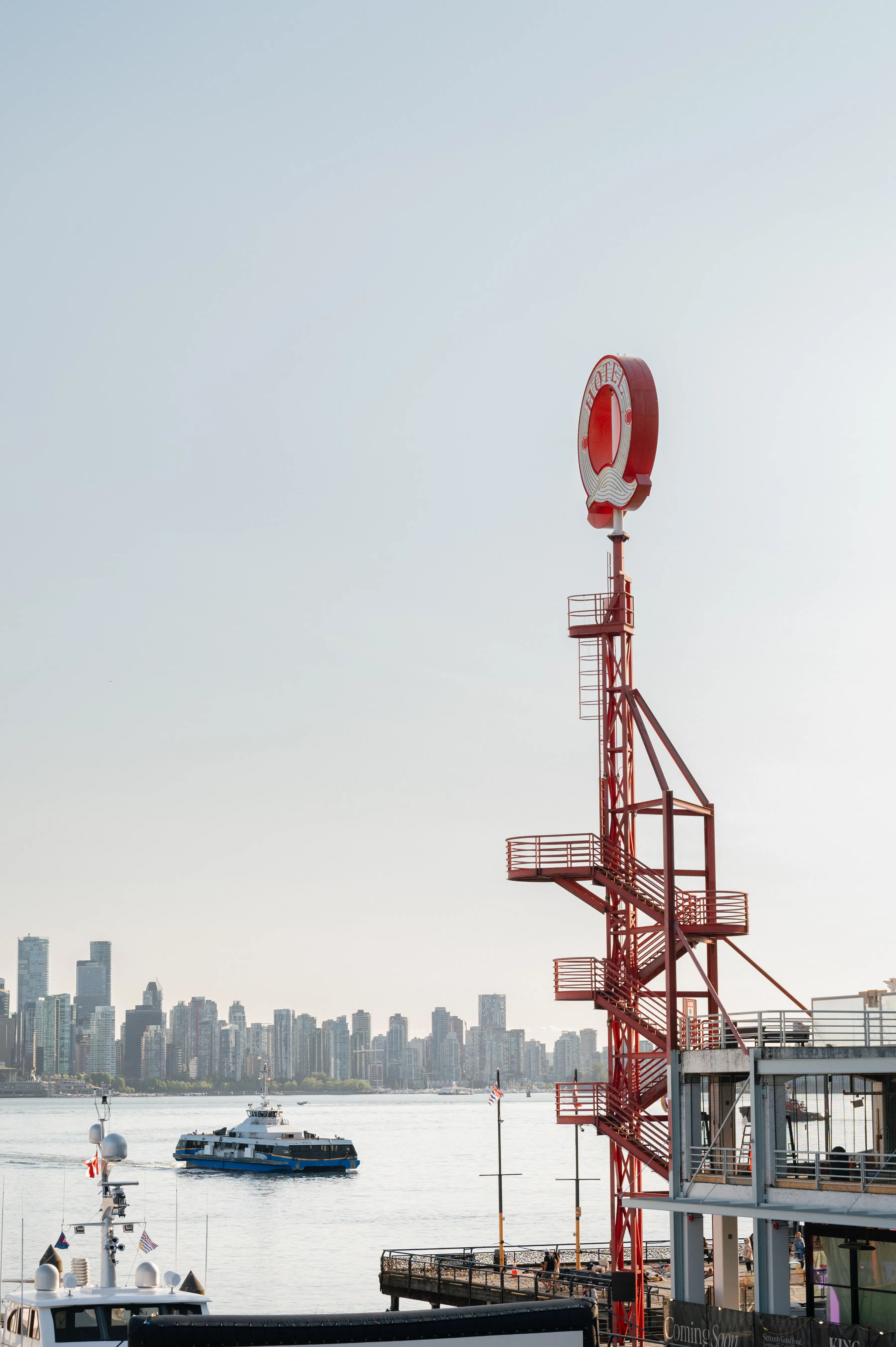 Red metal tower with a circular sign near a waterfront with boats and a city skyline in the background.