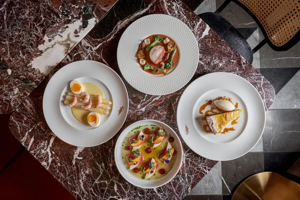 Four gourmet dishes on white plates arranged on a marbled table, including shrimp with eggs, a vegetable and seafood dish, a dessert with ice cream, and a colorful fruit and sauce dish.
