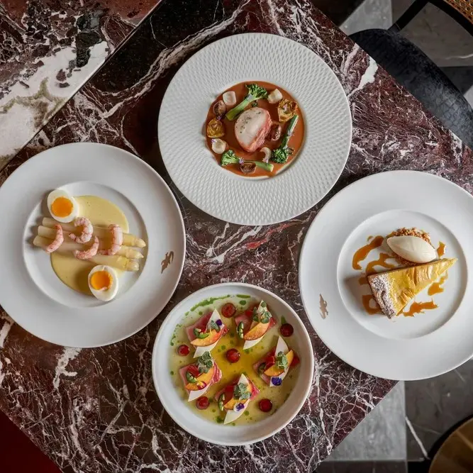 Four gourmet dishes on white plates arranged on a marbled table, including shrimp with eggs, a vegetable and seafood dish, a dessert with ice cream, and a colorful fruit and sauce dish.