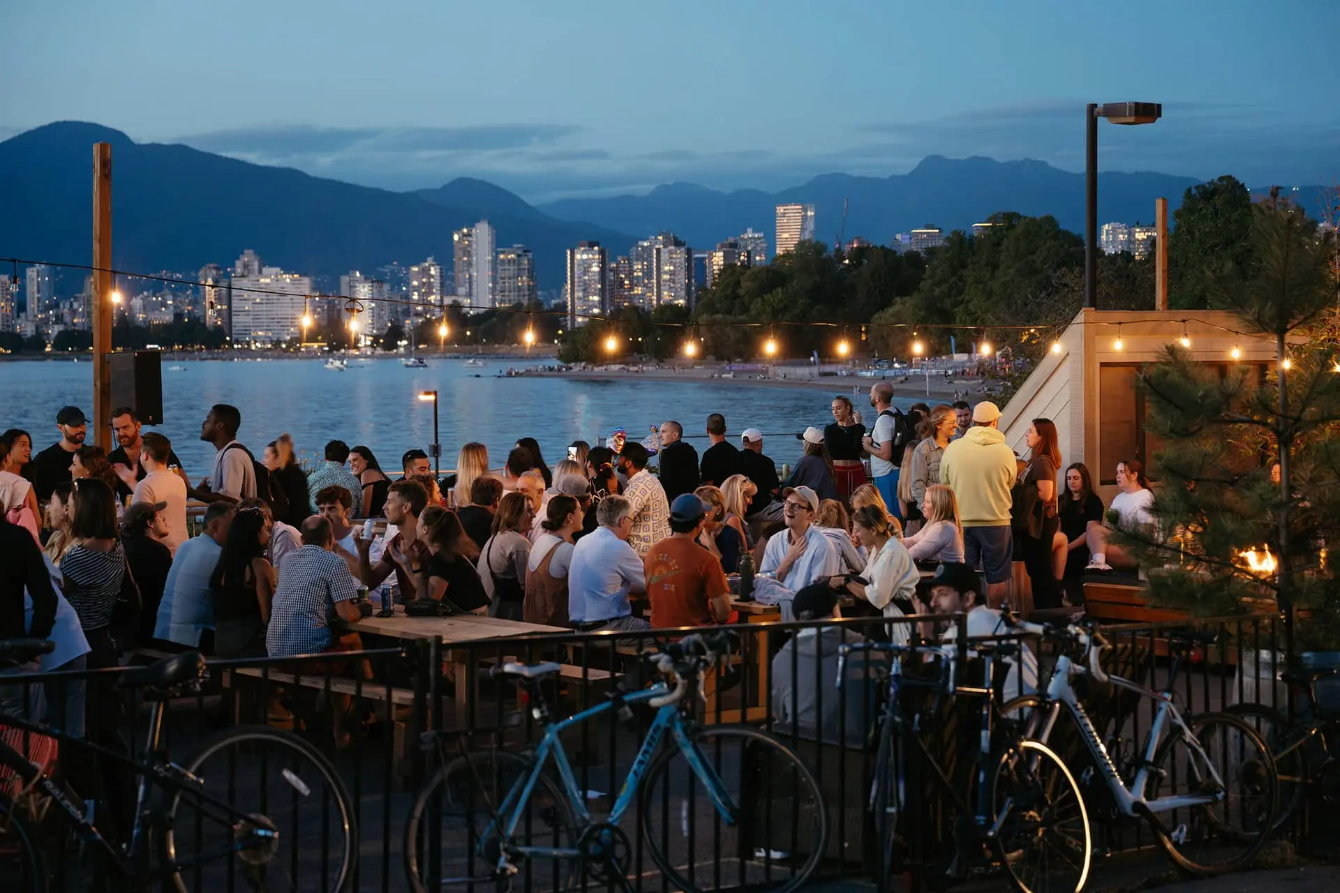 Crowd of people socializing at an outdoor waterfront venue with city skyline and mountains in the background at dusk.