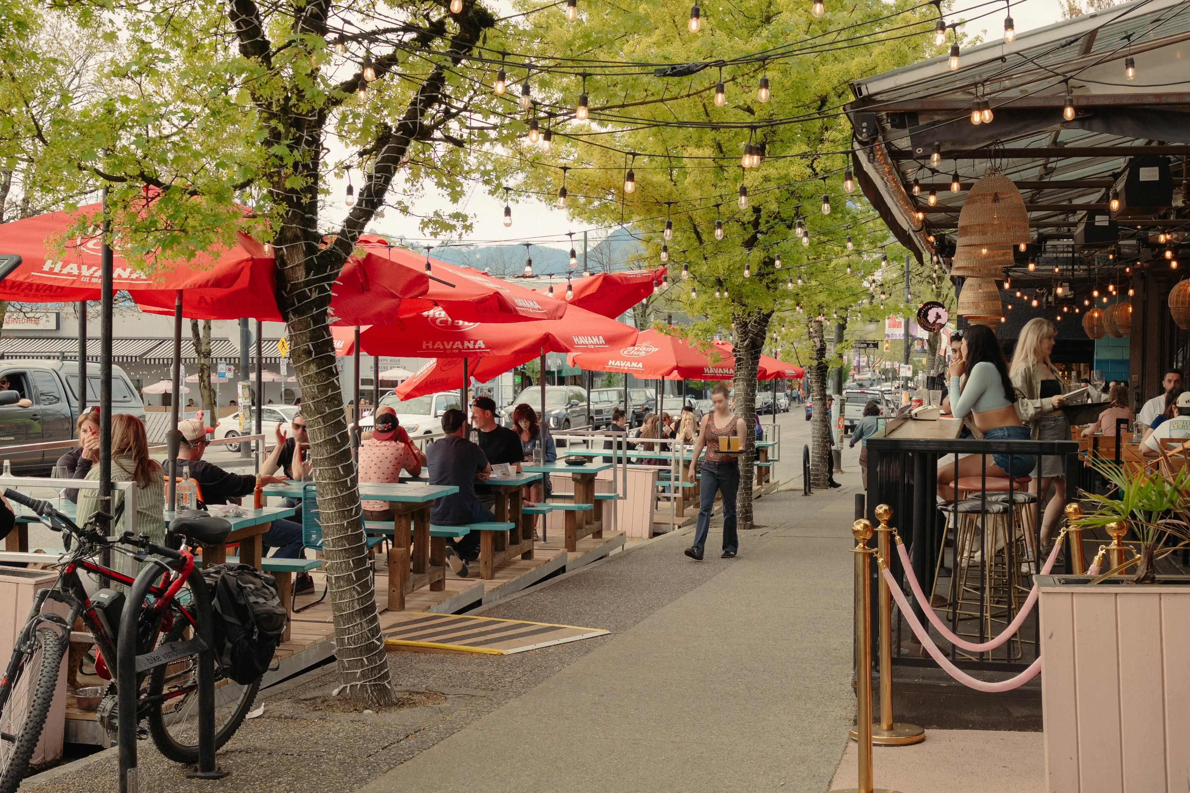 Outdoor dining area with people sitting under umbrellas and string lights, a waitress carrying drinks, and a person sitting at a bar at Havana in Vancouver.