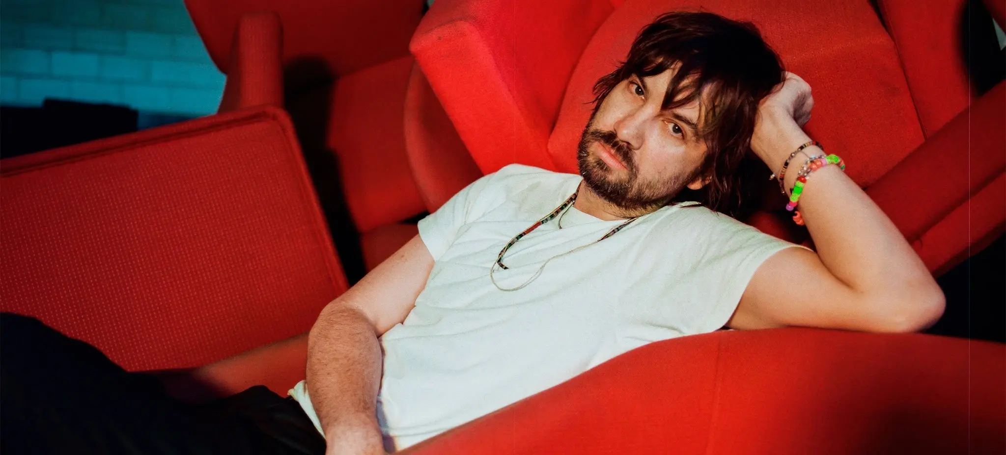 A person with dark hair and beard reclining on red chairs, wearing a white t-shirt and colorful bracelets.