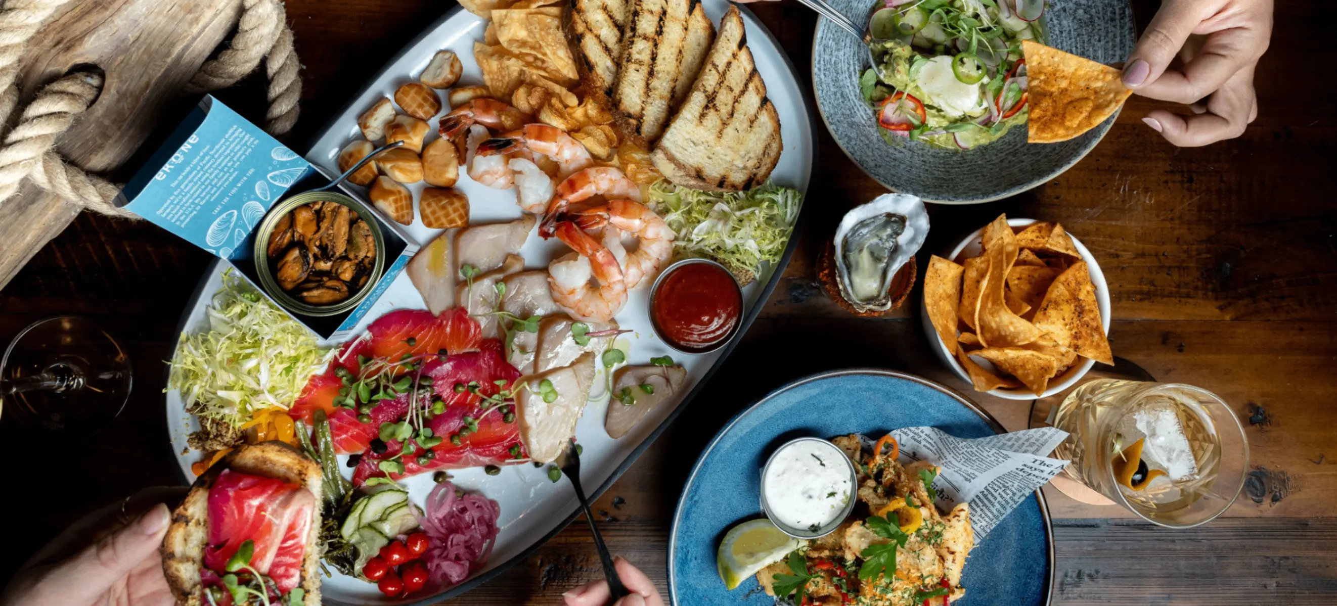 Seafood platter with shrimp, smoked fish, grilled bread, and sauces, with hands holding a chip and a piece of bread with fish.