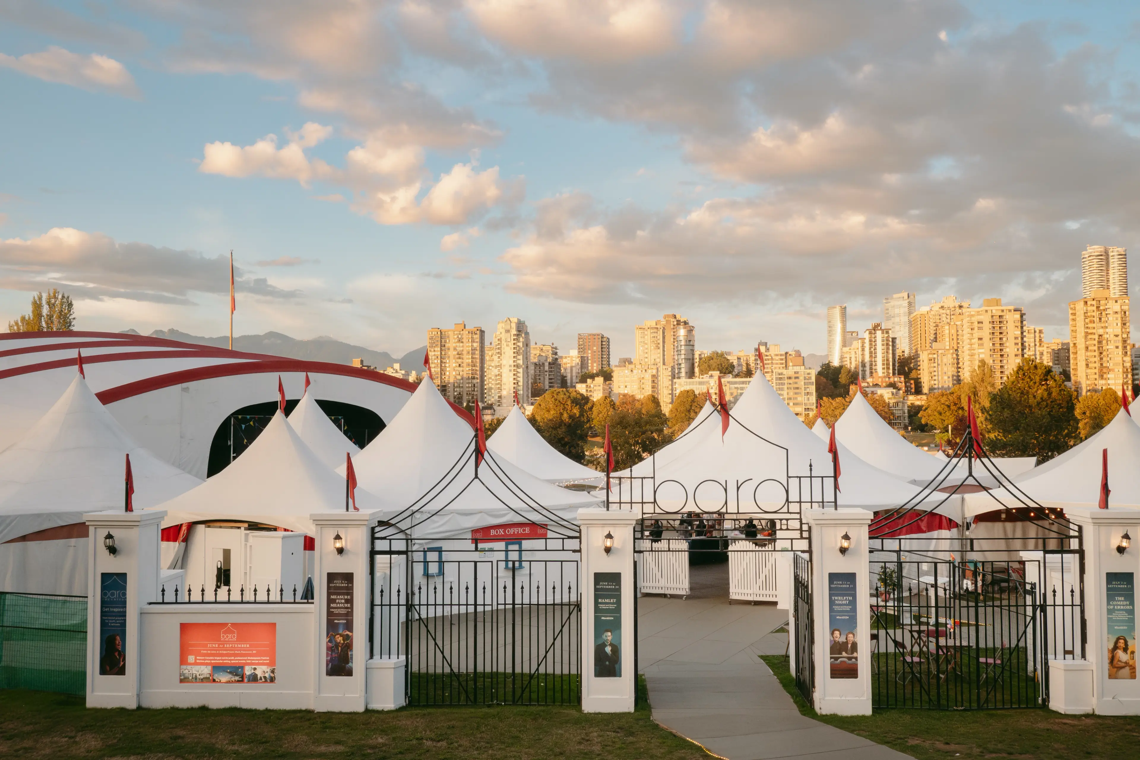 White tents with red flags and a black iron gate entrance labeled 'bard' in front of a city skyline under a partly cloudy sky.