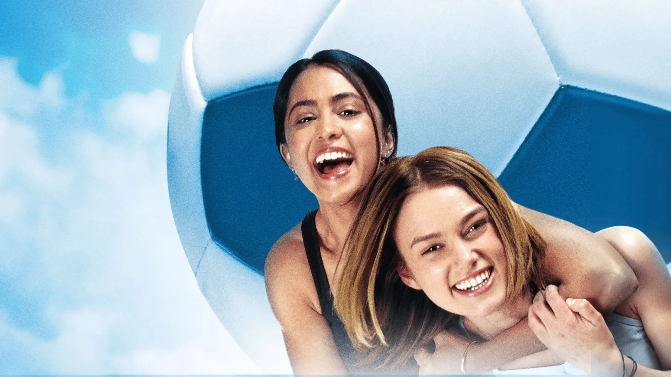 Two people smiling and hugging in front of a large soccer ball with a blue sky background.