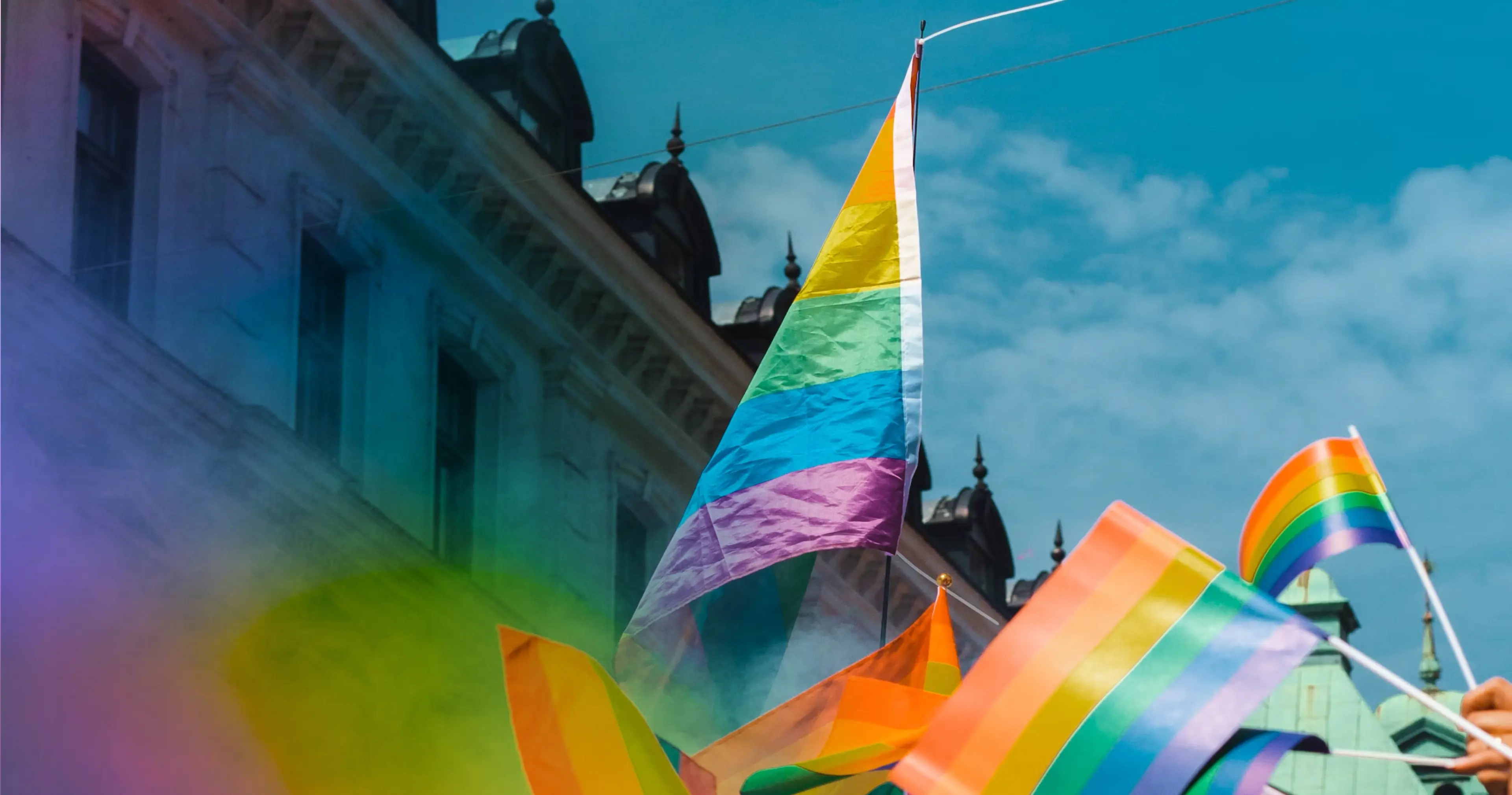 A rainbow Pride flag waves in the air among several others during a sunny day in Vancouver city, with a historic building and blue sky in the background.