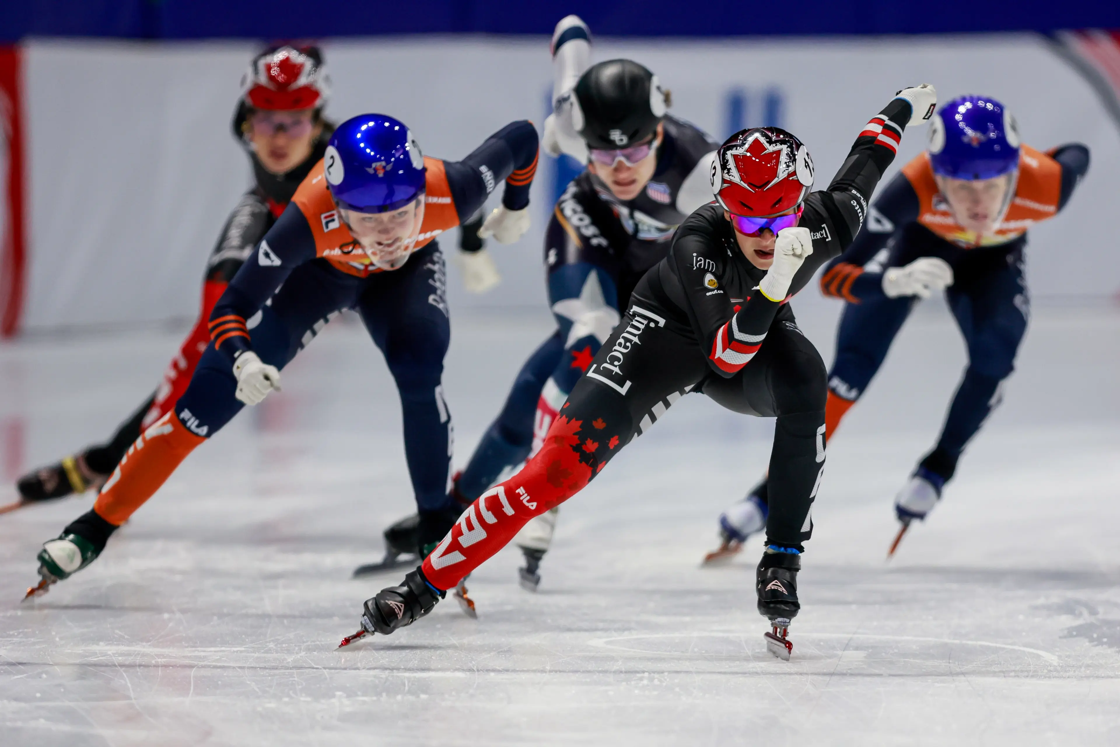 Five speed skaters racing on ice, with the leading skater wearing a black and red suit and helmet with a maple leaf design.