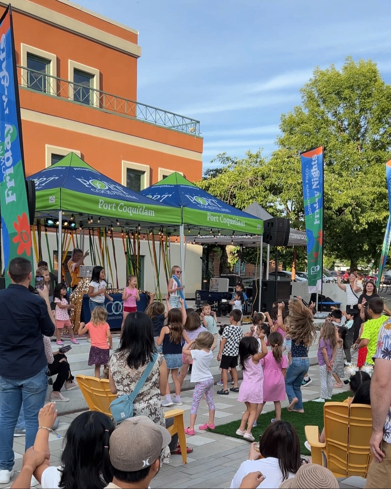 Children dancing and playing in front of a stage with green tents labeled Port Coquitlam during an outdoor event with adults watching.