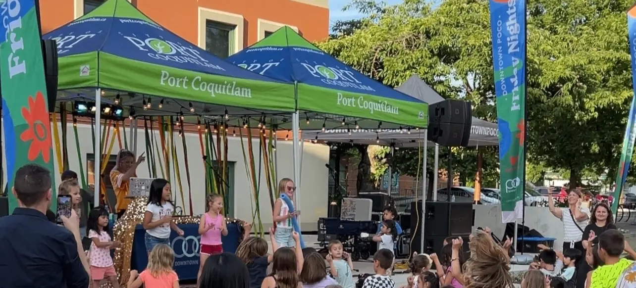 Children dancing and playing in front of a stage with green tents labeled Port Coquitlam during an outdoor event with adults watching.