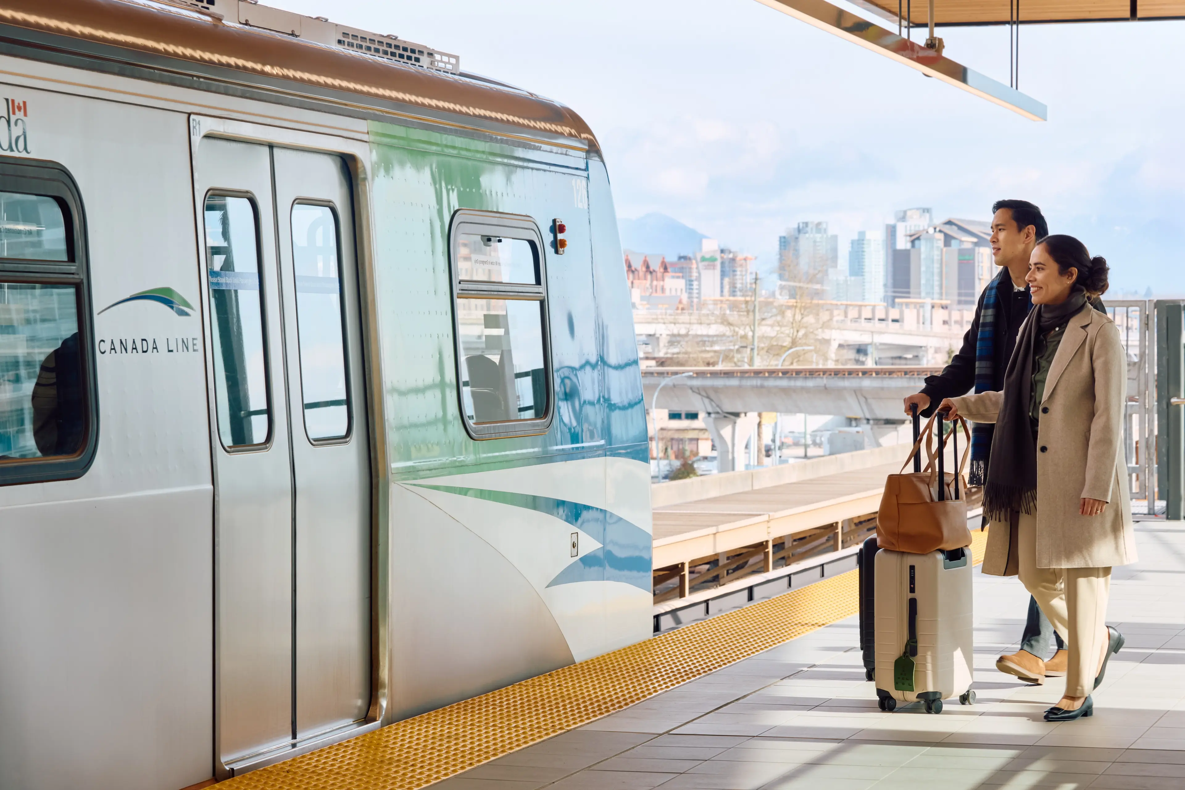Two travellers board the SkyTrain with suitcases, with Vancouver’s skyline and mountains in the background.