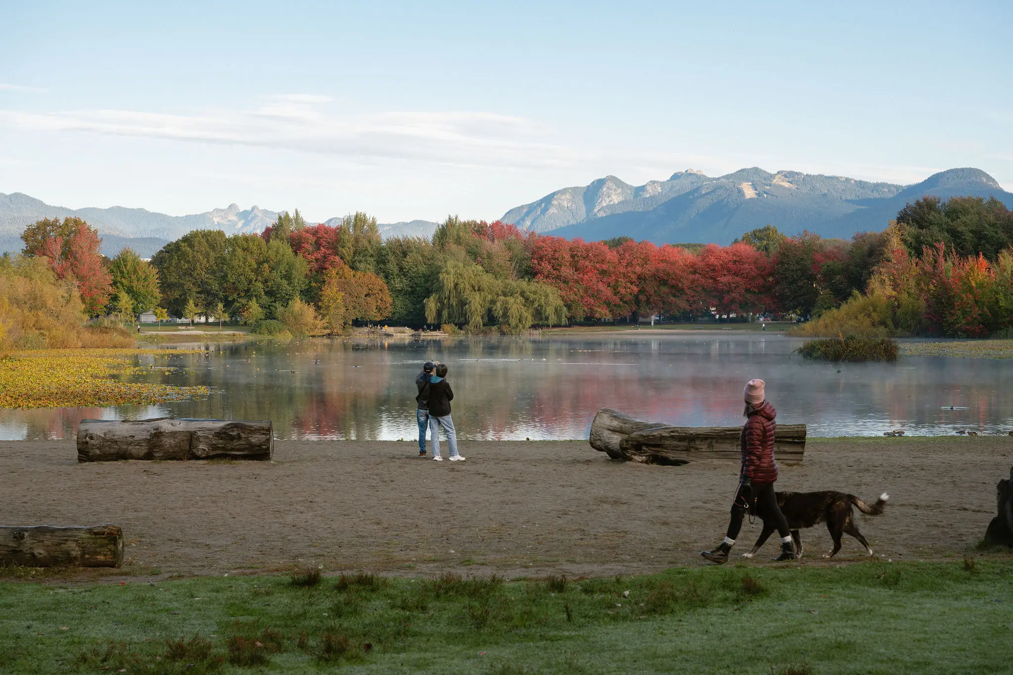 Person walking a dog on a sandy path by a lake with autumn trees and mountains in the background.