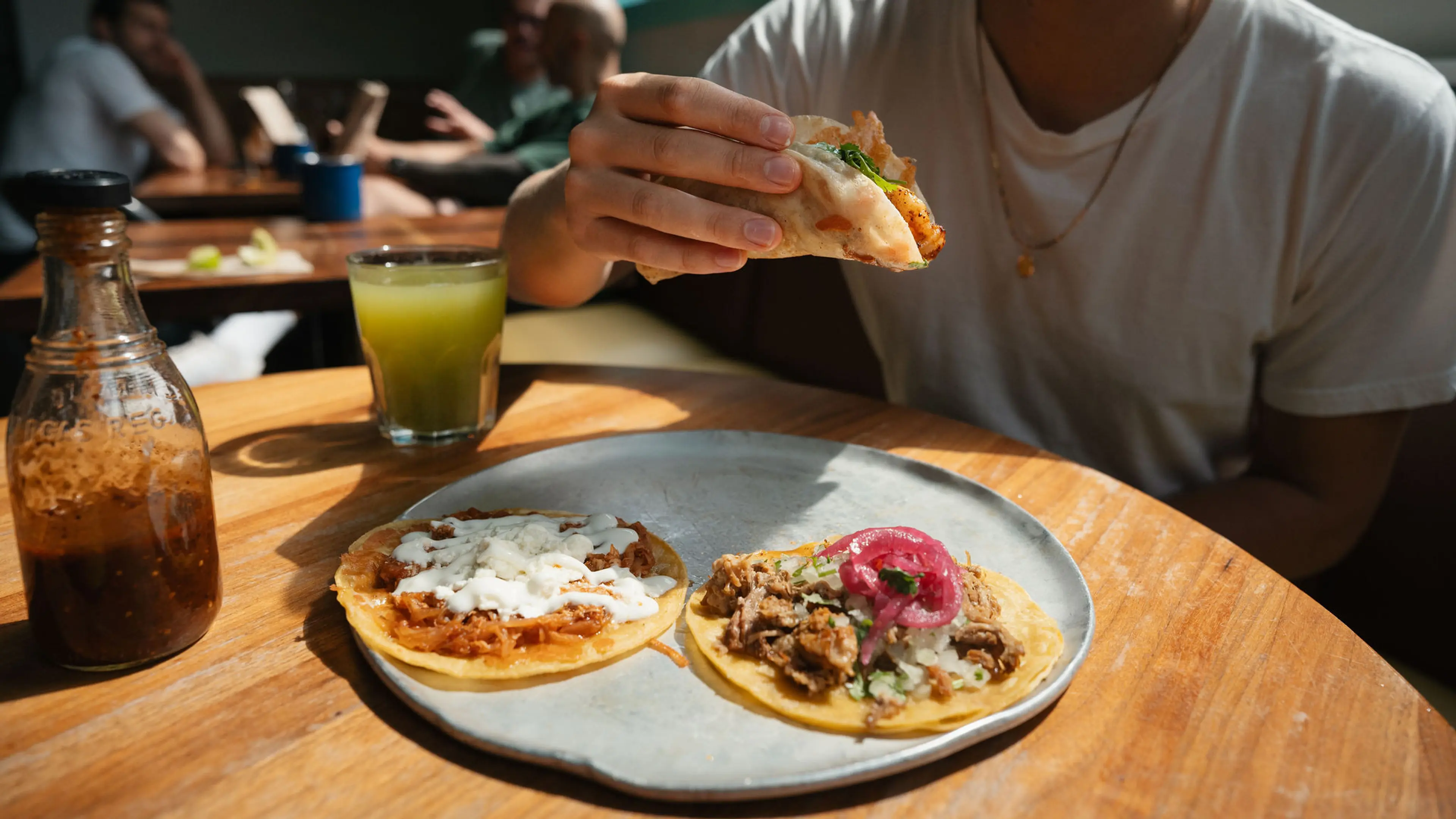 Person holding a taco over a plate with two tacos on a wooden table, with a glass of green drink and a bottle of sauce nearby.