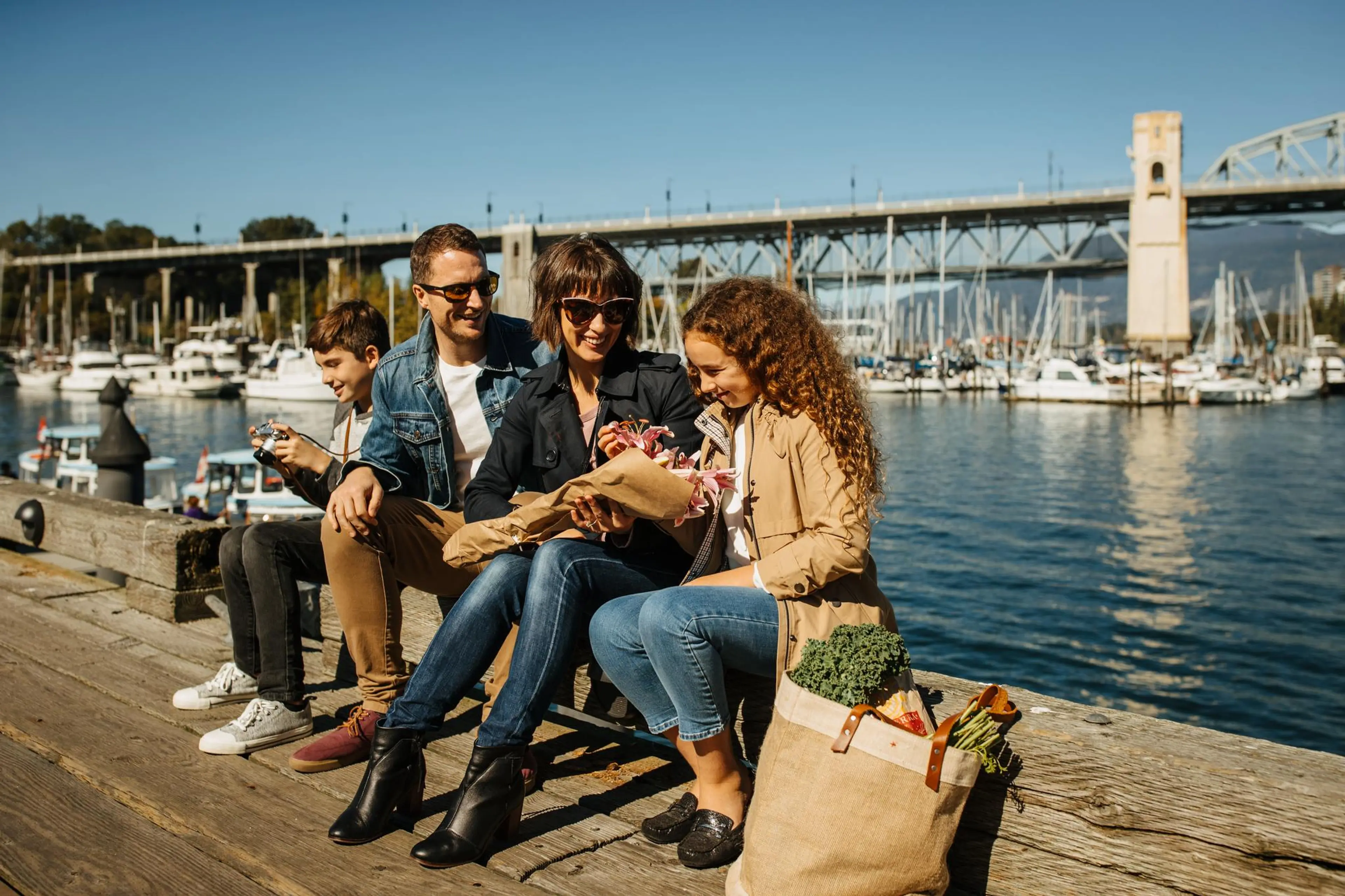 Family of four sitting on a dock by the water with boats and a bridge in the background, woman holding flowers, grocery bag nearby.