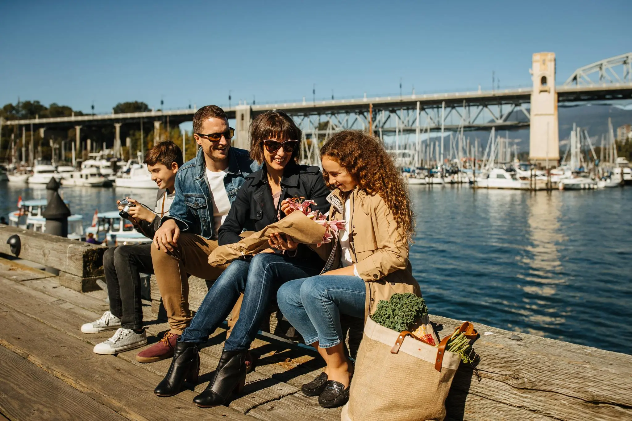 Family of four sitting on a dock by the water with boats and a bridge in the background, woman holding flowers, grocery bag nearby.