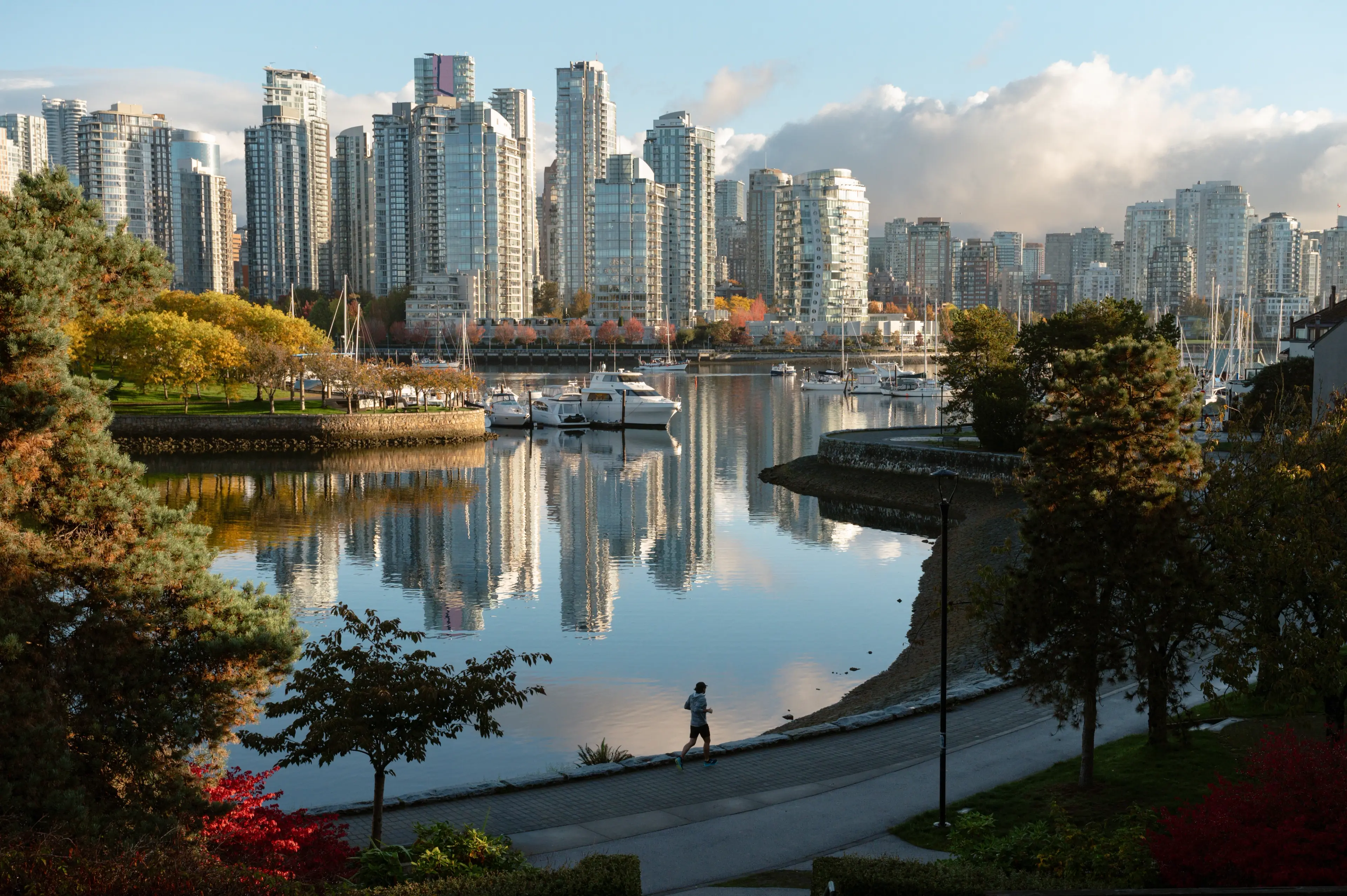 Person running along the seawall in False Creek with the downtown Vancouver skyline in the background, framed by trees in vibrant fall colours beside the water.