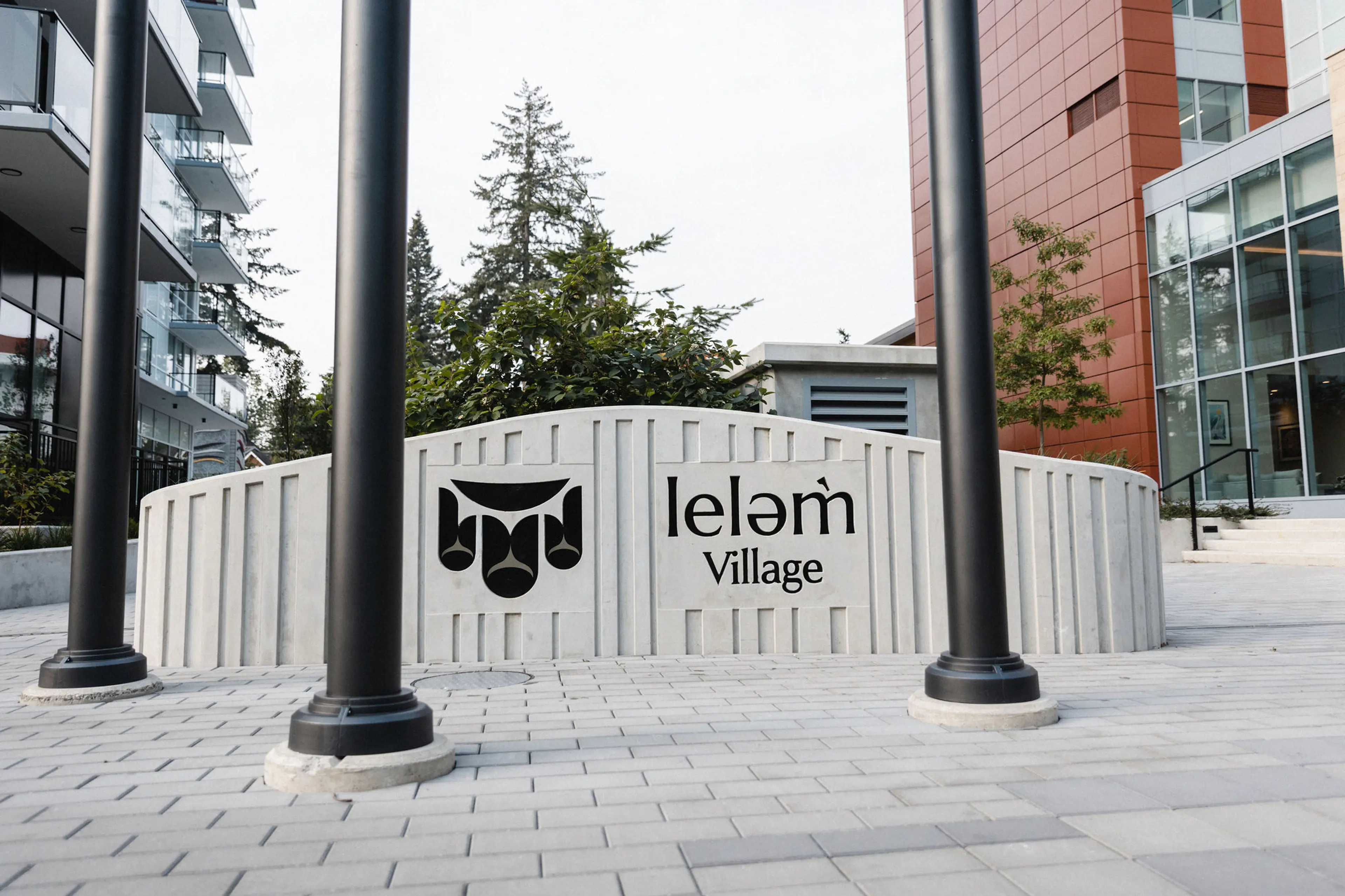 Concrete sign with black logo and text reading 'lelem Village' between three black poles on a paved area with buildings and trees in the background.