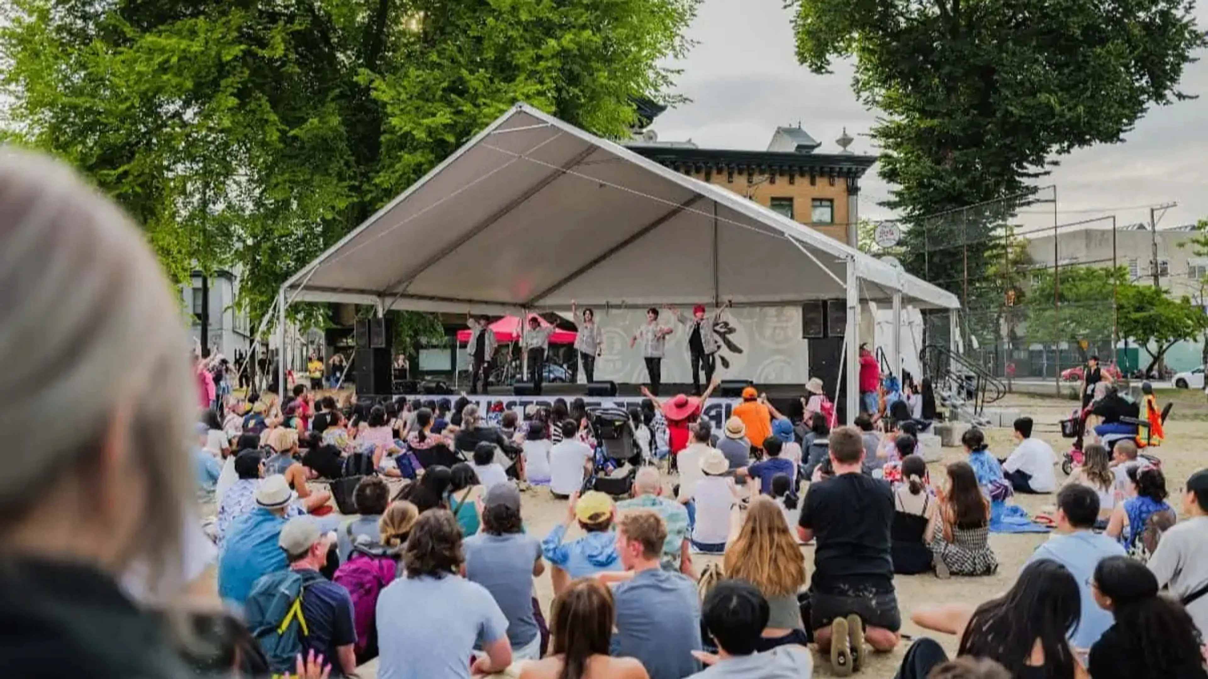 A crowd sitting on the ground watching a group of performers on a stage under a white tent outdoors.