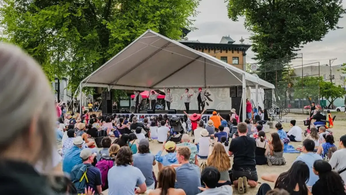 A crowd sitting on the ground watching a group of performers on a stage under a white tent outdoors.
