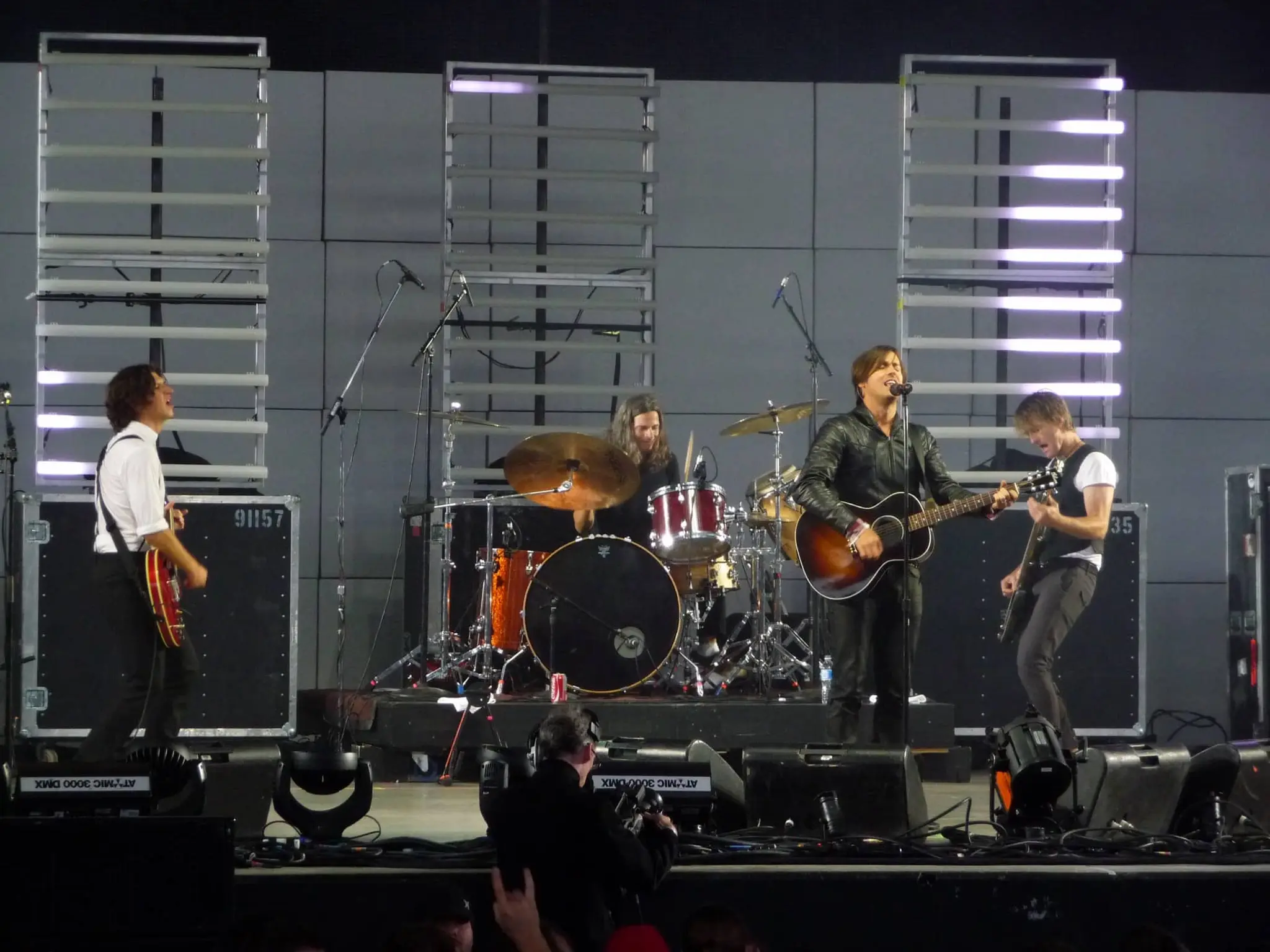 Four-member band performing on stage with guitars and drums, illuminated by vertical light panels.