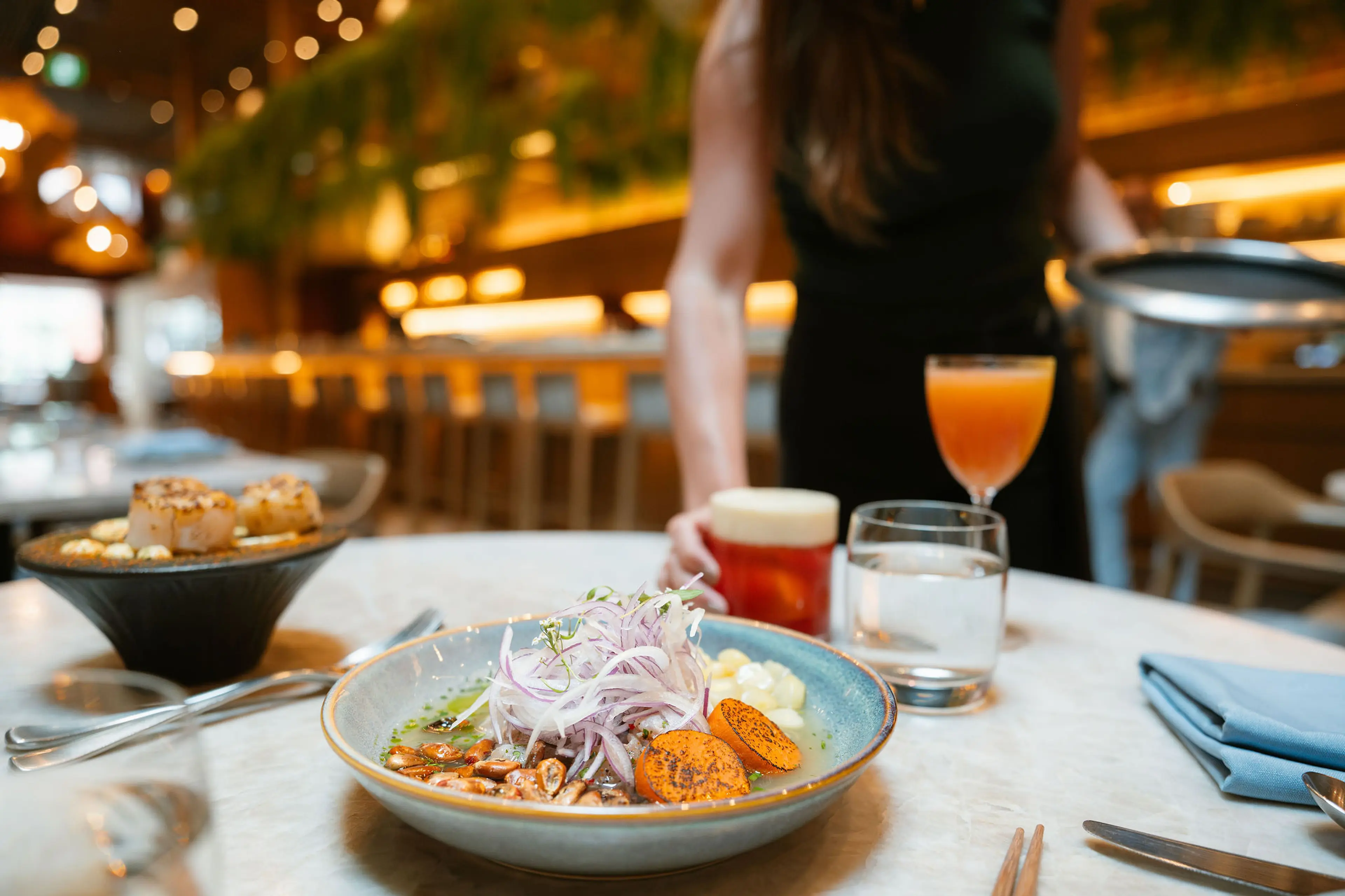 A bowl of food with sliced onions, corn, and orange slices on a table with drinks and a person in the background in a restaurant.