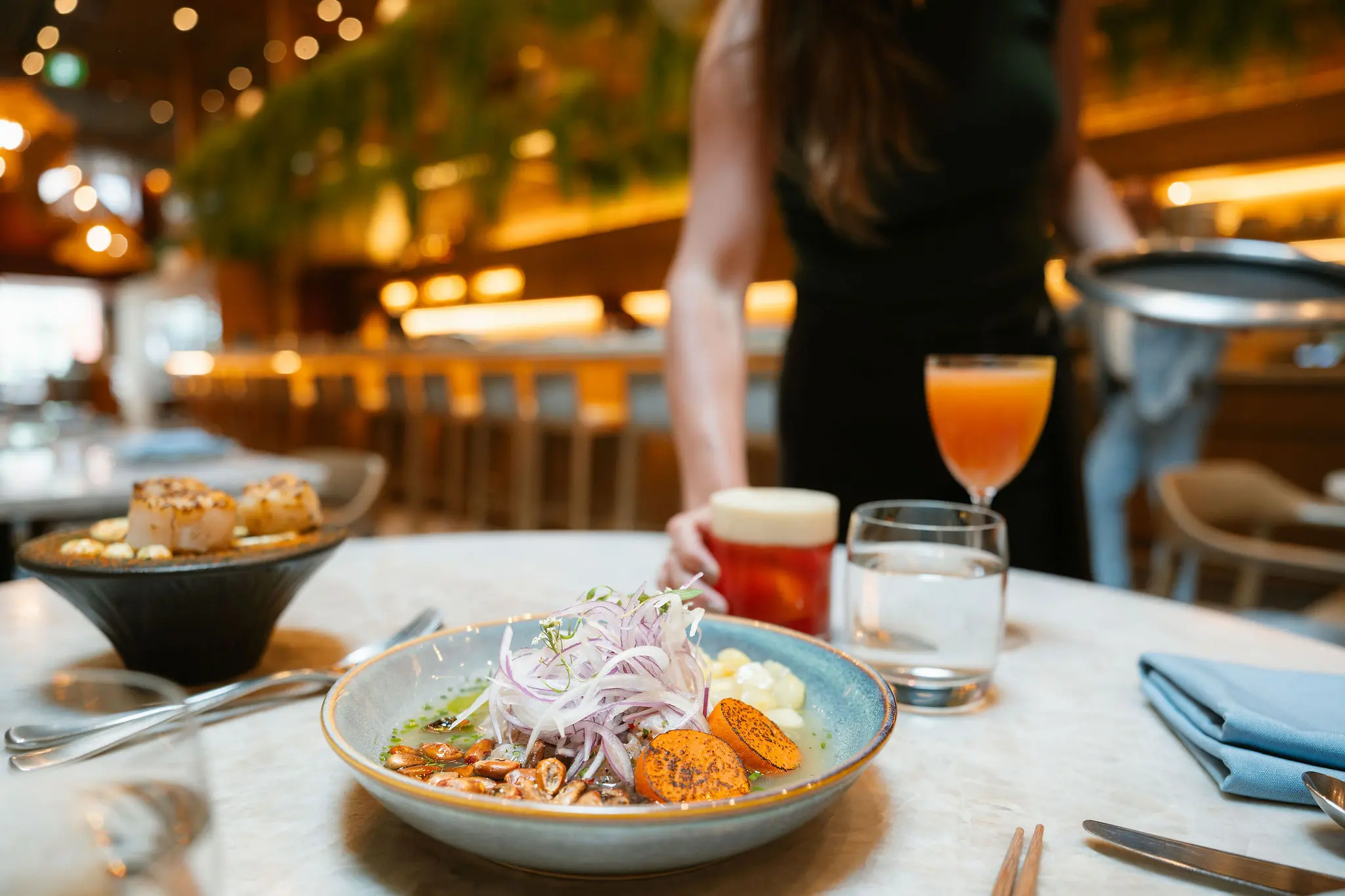 A bowl of food with sliced onions, corn, and orange slices on a table with drinks and a person in the background in a restaurant.