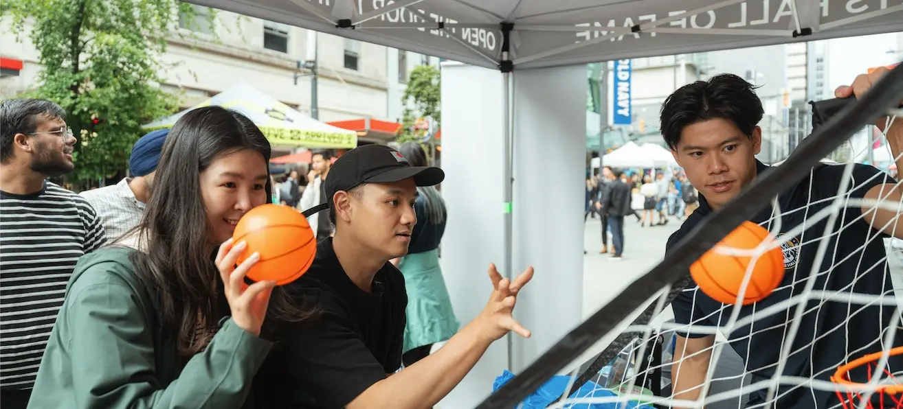 Three people playing an arcade basketball game under a tent, one person holding an orange ball ready to throw it.