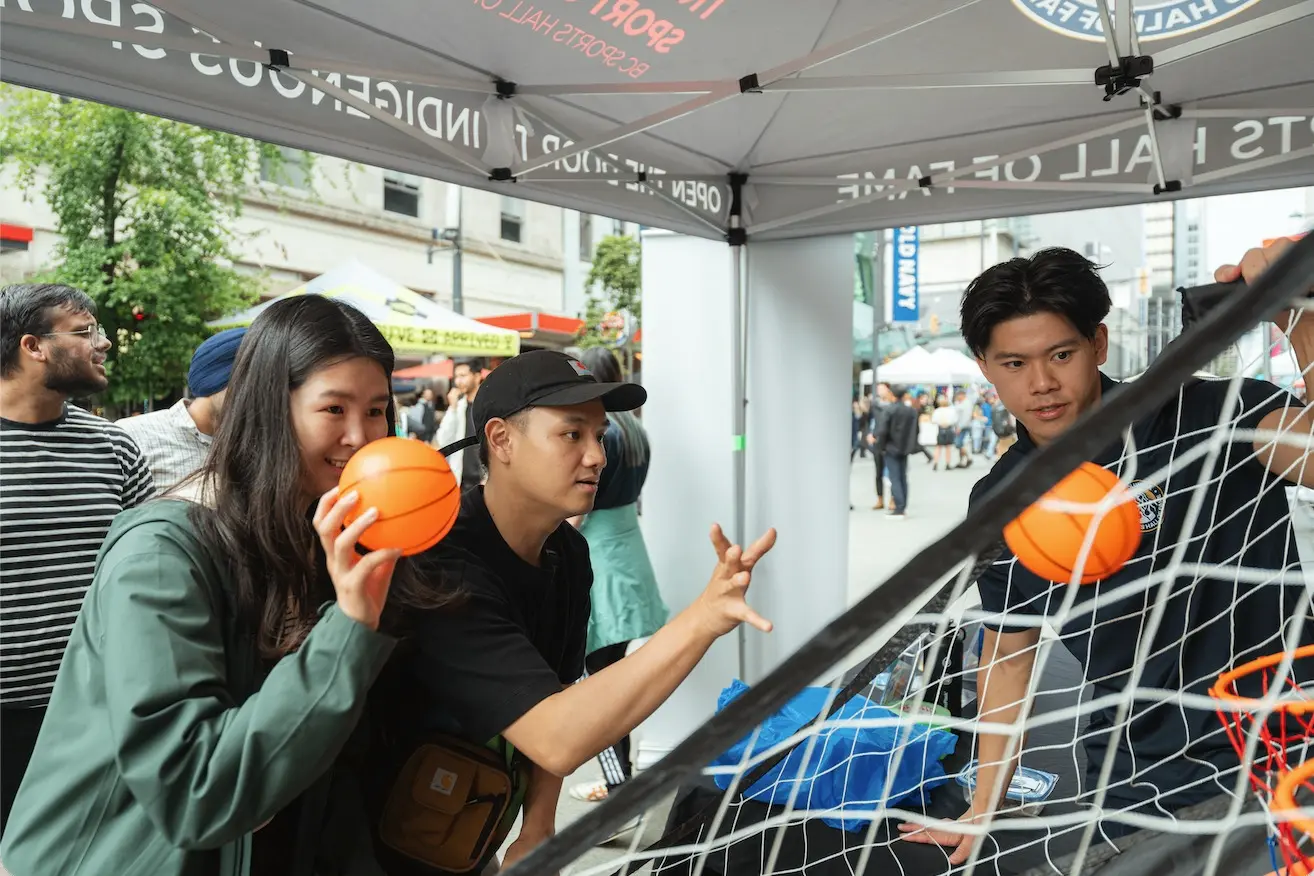 Three people playing an arcade basketball game under a tent, one person holding an orange ball ready to throw it.