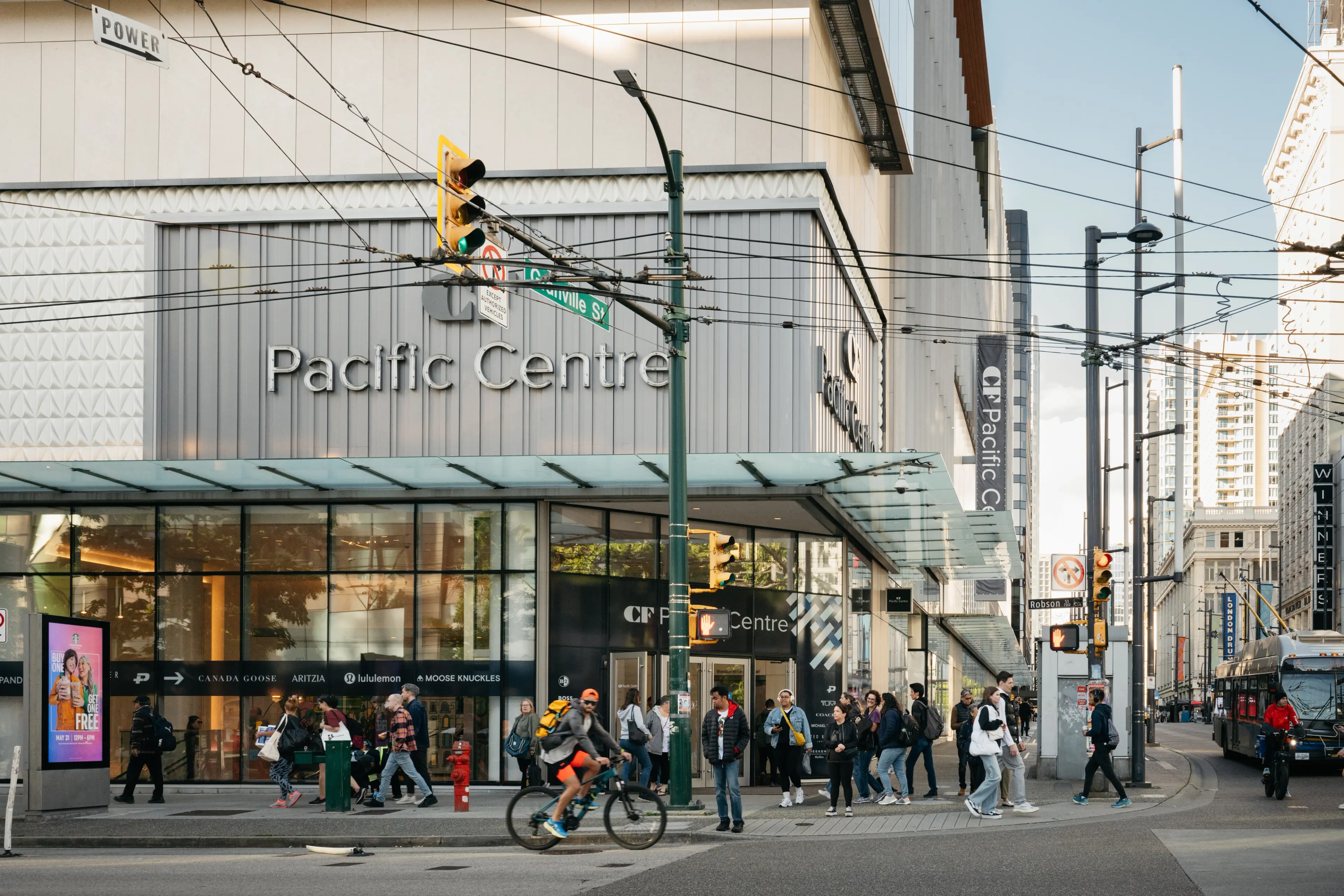 People walking and a cyclist in front of the Pacific Centre shopping mall on a city street with traffic lights and overhead wires.