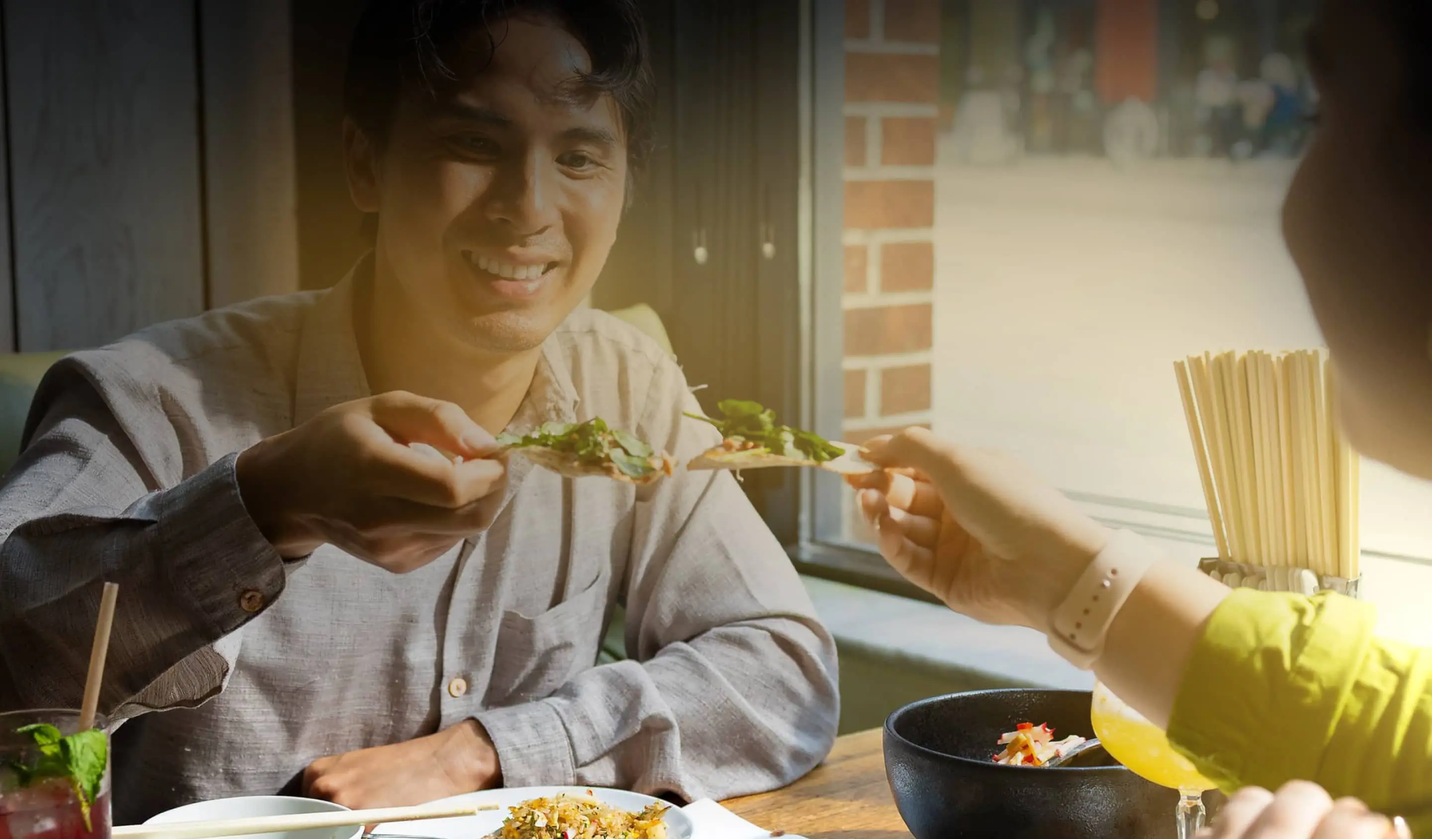 Two people clinking slices of pizza at a restaurant table near a window with sunlight.