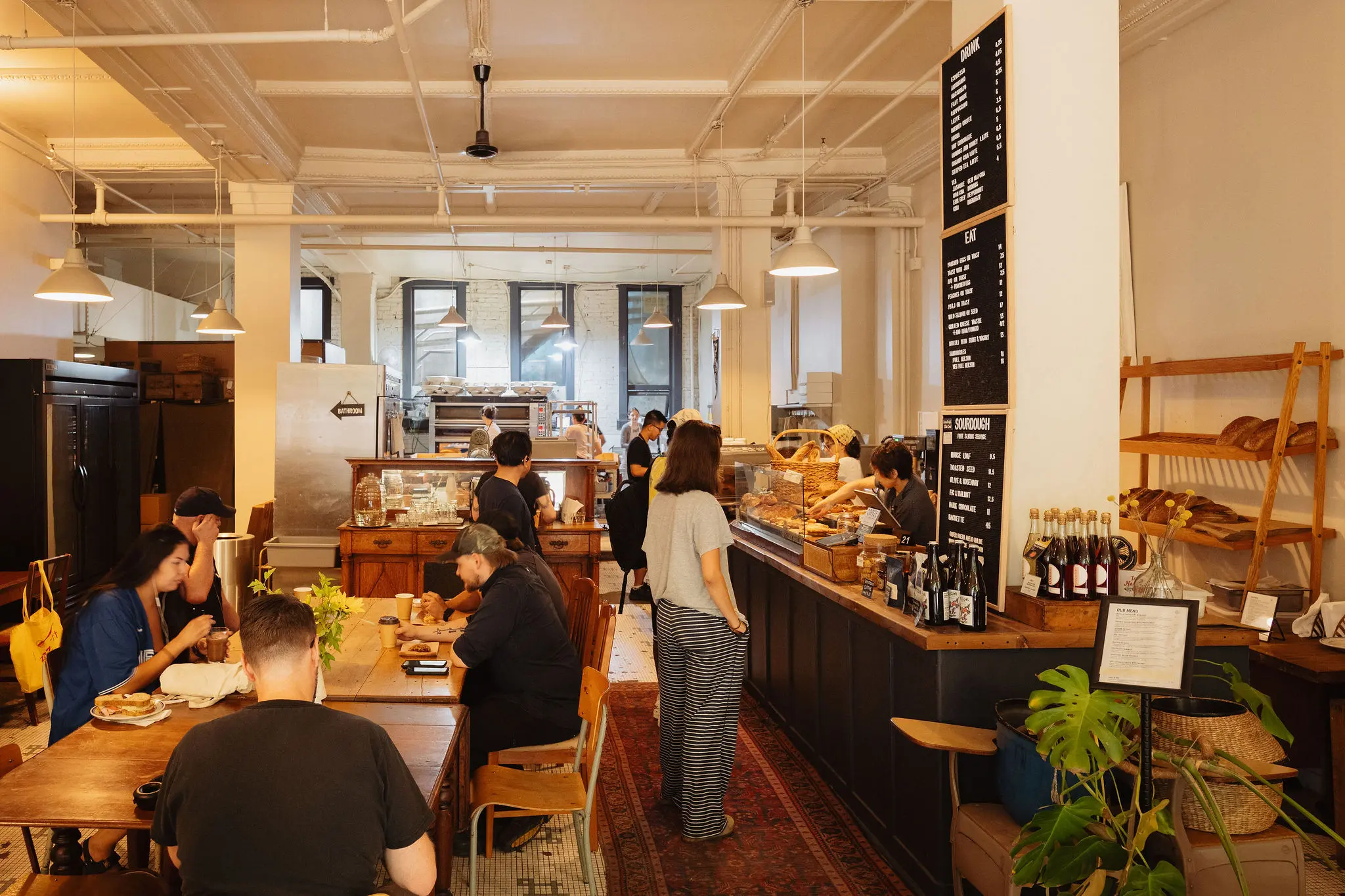 People sitting and standing inside a cozy bakery cafe with wooden tables, shelves of bread, and a menu on the wall.