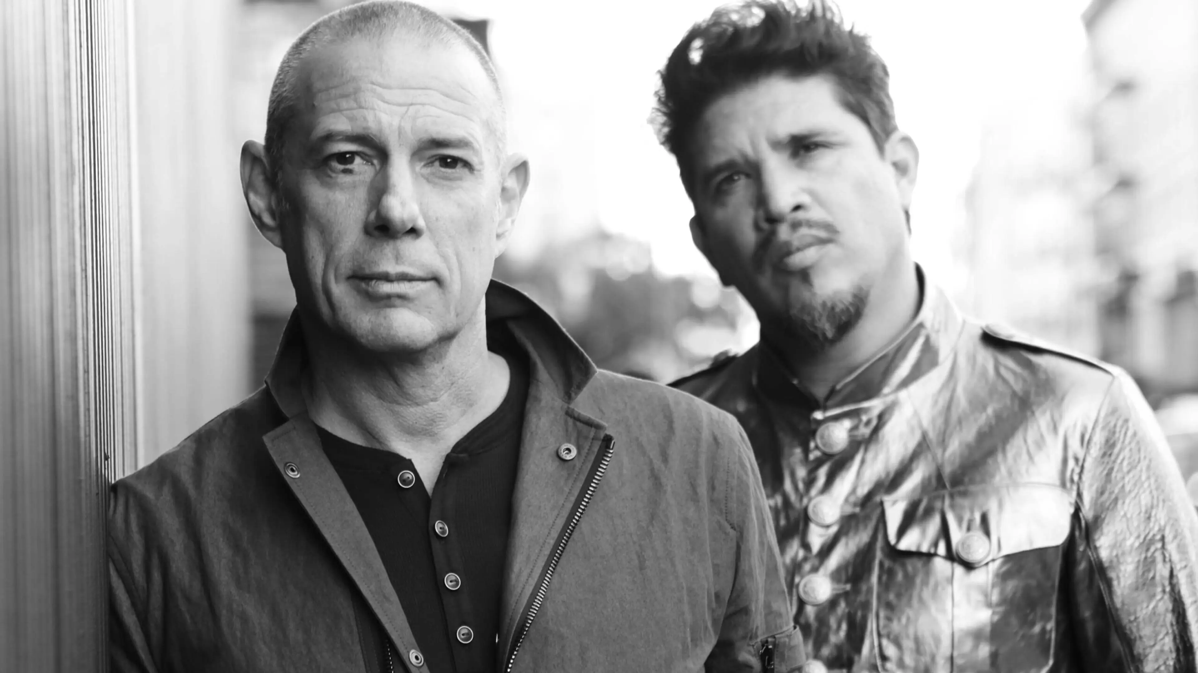 Two people standing outdoors, one with a shaved head wearing a jacket, the other with styled hair wearing a shiny buttoned jacket.