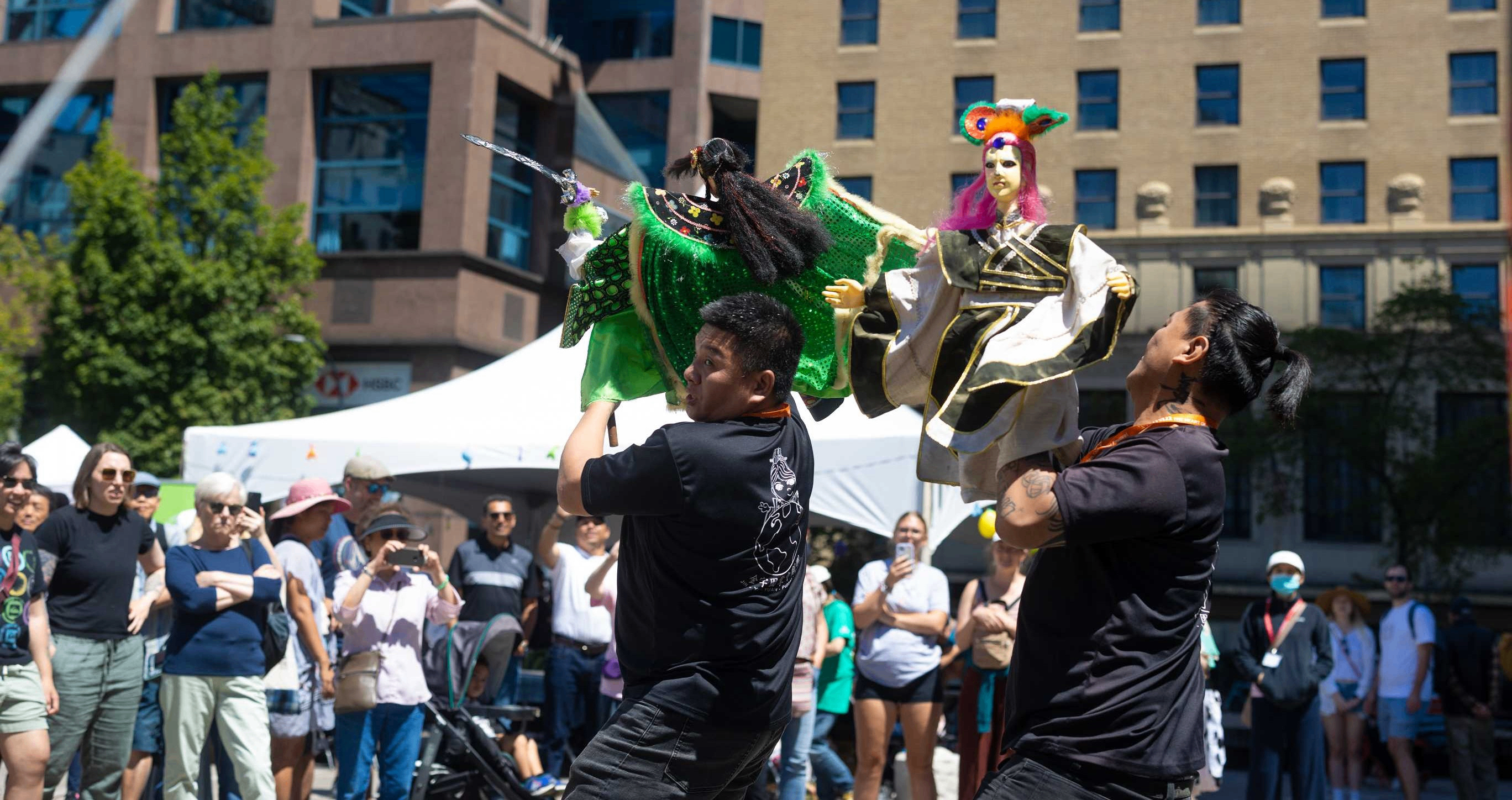 Two people holding up colorful puppets in front of a crowd at an outdoor event in a city plaza.