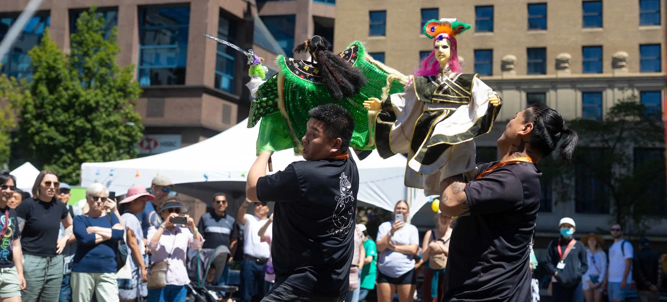 Two people holding up colorful puppets in front of a crowd at an outdoor event in a city plaza.
