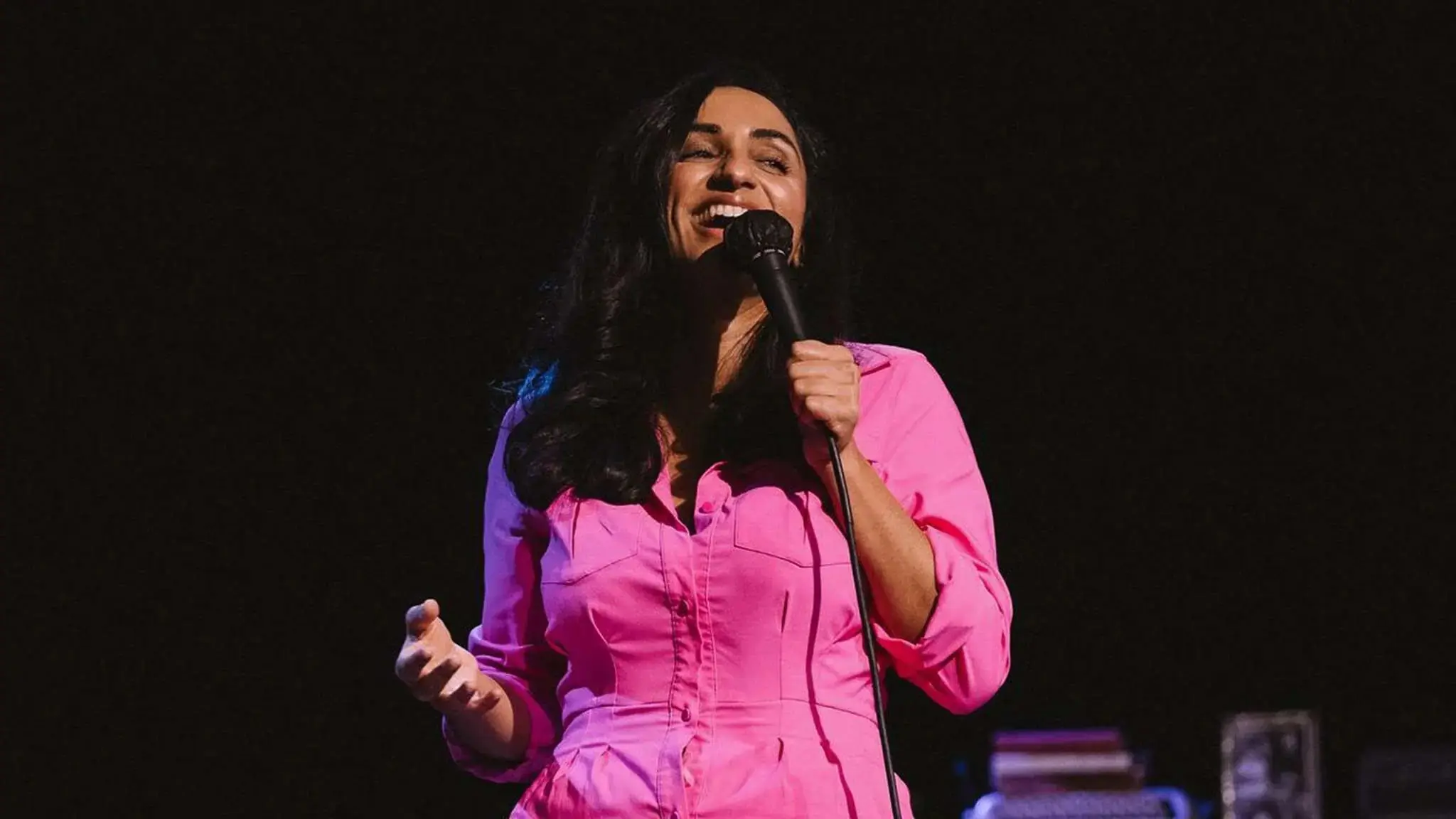 Person in a bright pink shirt holding a microphone and smiling on a dark stage.