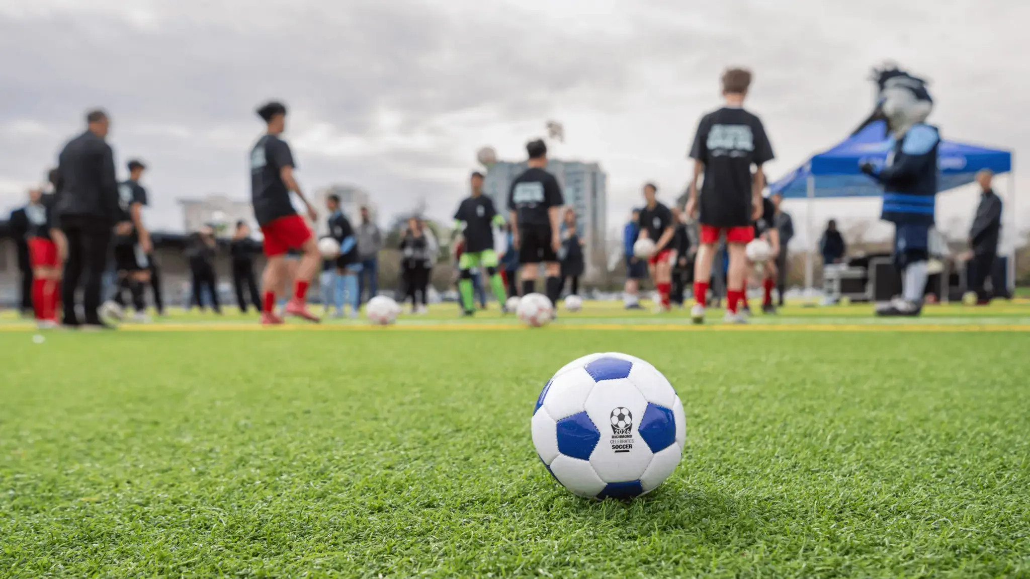 Soccer ball on grass with players and a mascot in the background on a cloudy day.