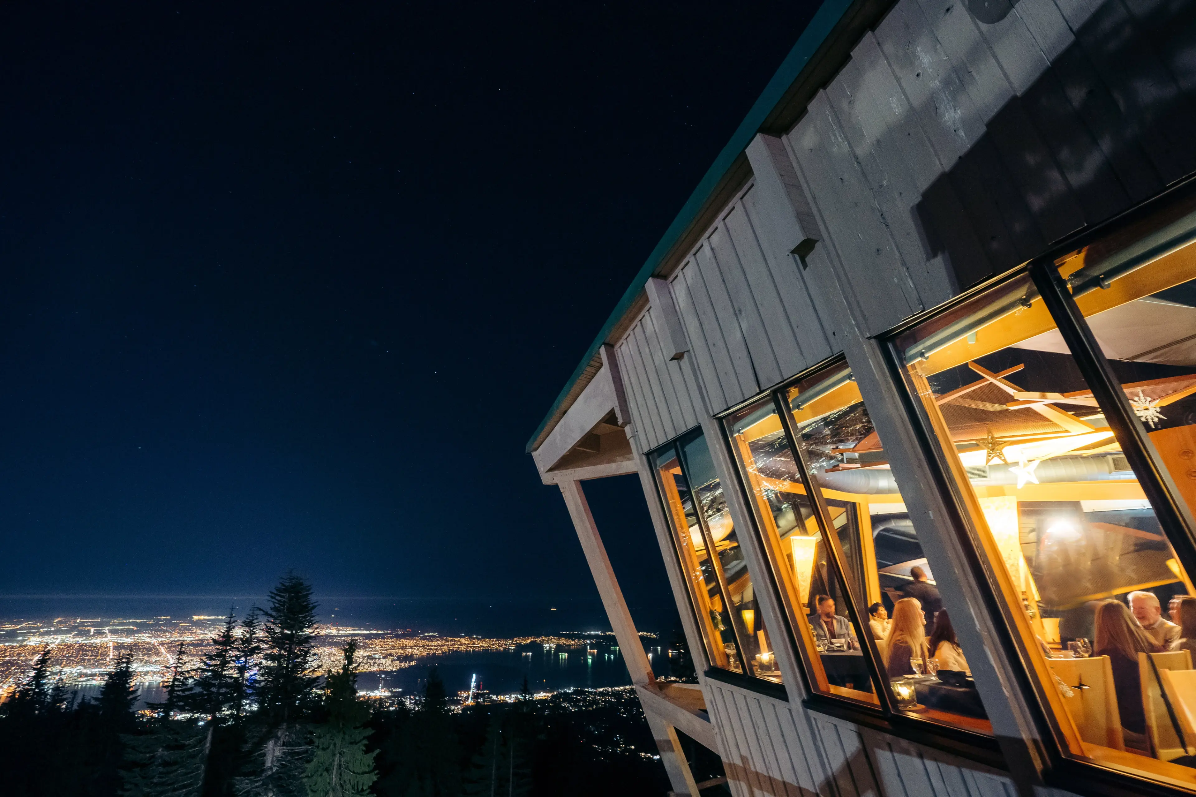 The exterior of the Observatory Restaurant on Grouse Mountain. You can see people dining through the window and the city light in the distance. 