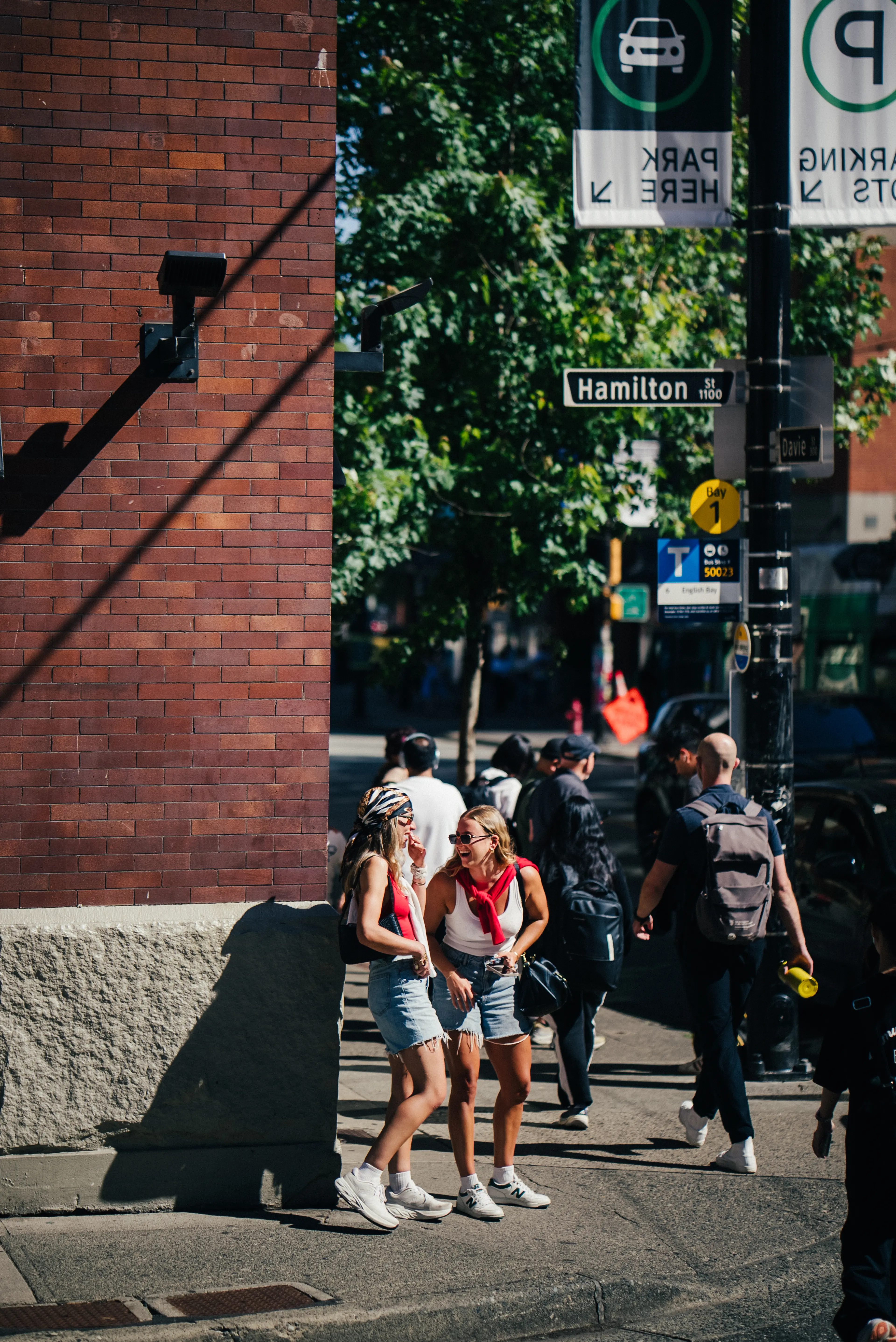 Two women in casual summer clothes laughing and talking on a sunny city street corner near a Hamilton St sign.