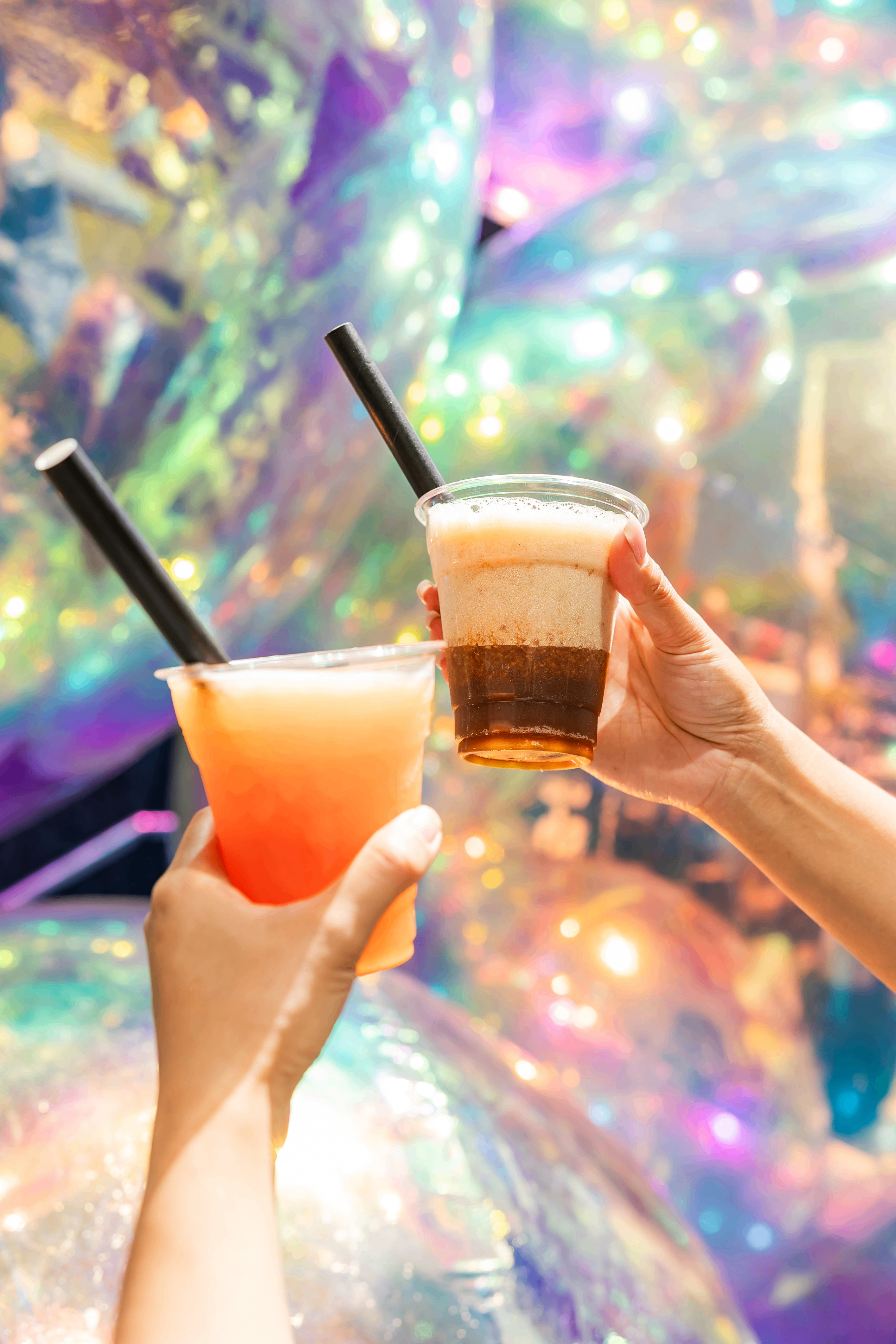 Two hands holding plastic cups with bubble tea drinks and black straws against a colorful, iridescent background.