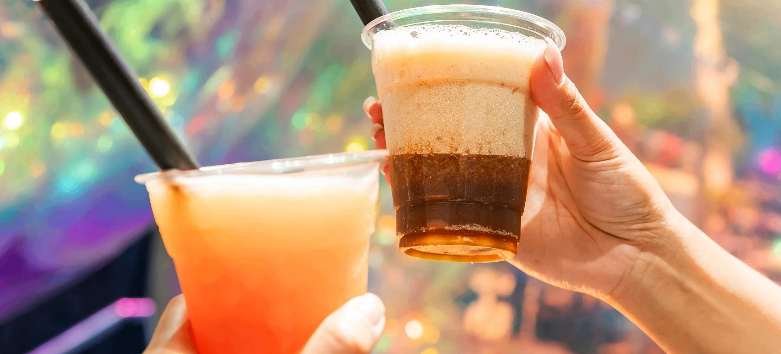 Two hands holding plastic cups with bubble tea drinks and black straws against a colorful, iridescent background.