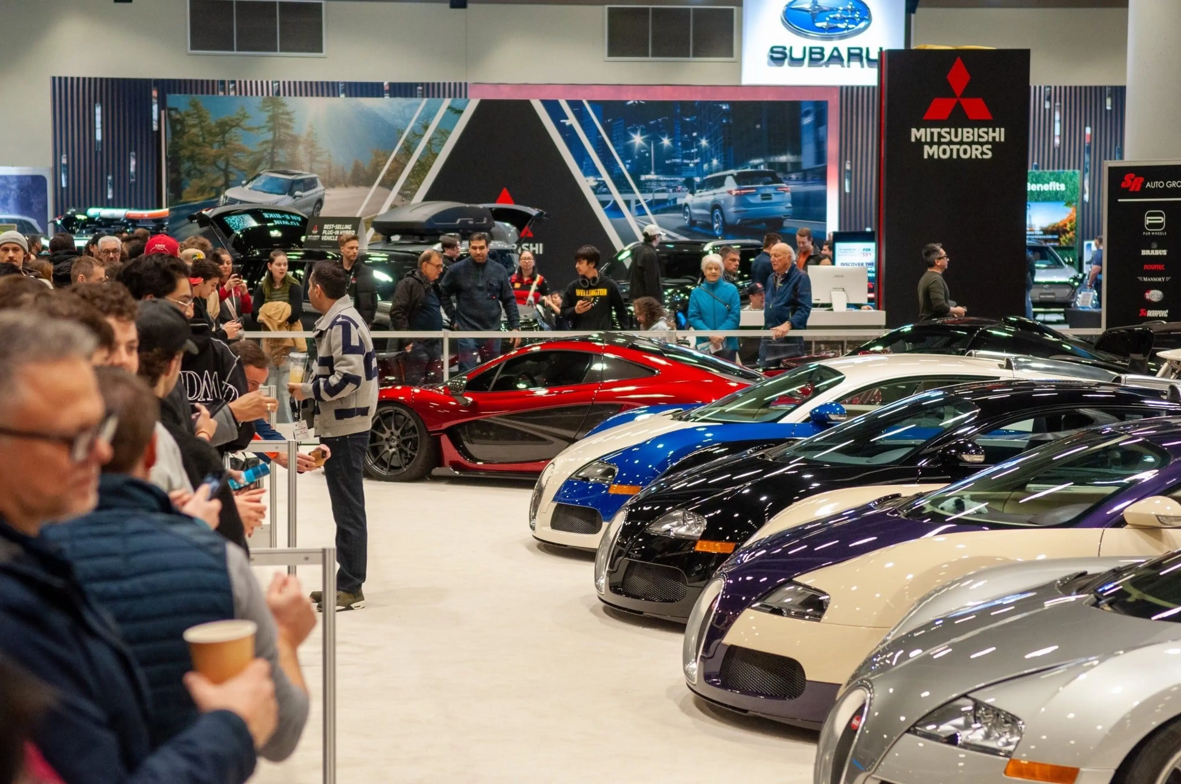 Crowds standing in line to view a row of luxury cars at the Vancouver International Auto Show.