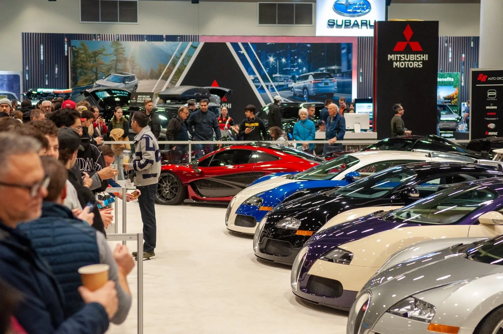Crowds standing in line to view a row of luxury cars at the Vancouver International Auto Show.