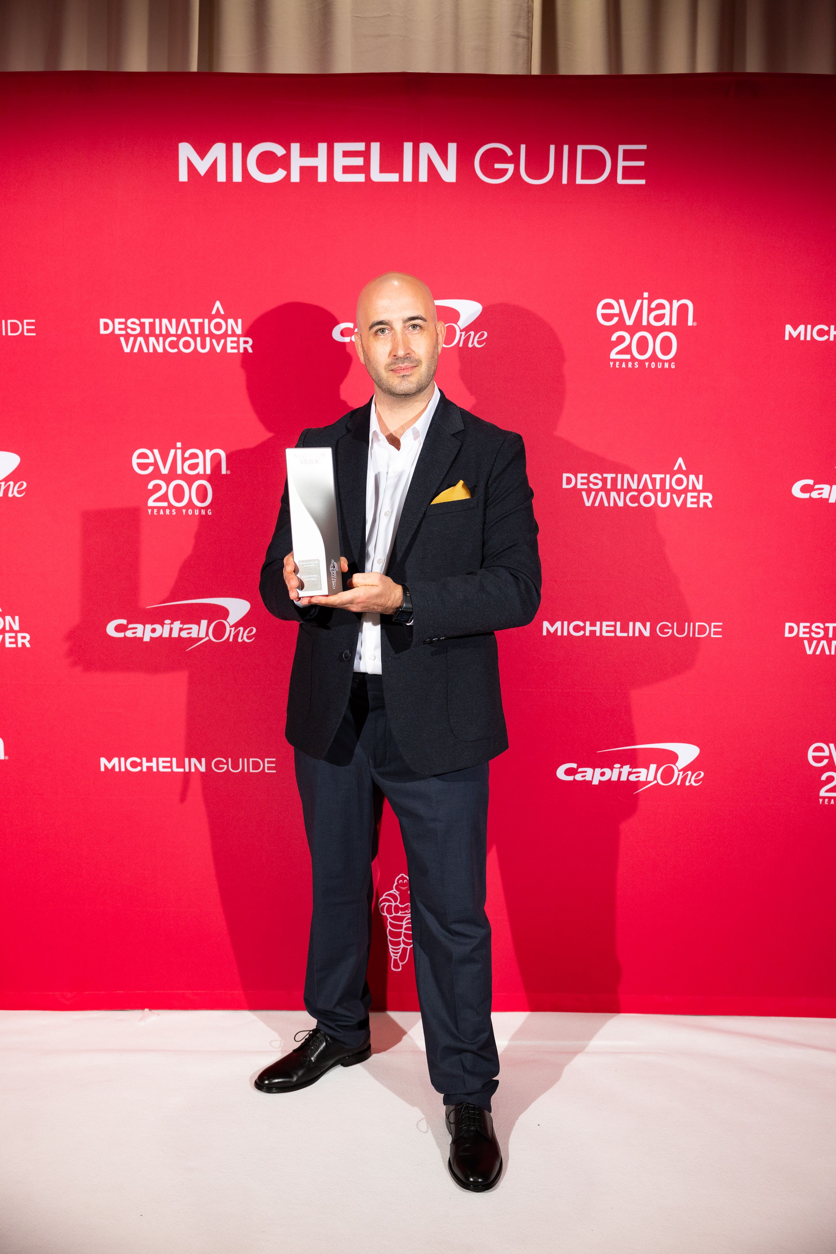 Chris Enns, the winner of the MICHELIN Exceptional Cocktails award, in a black suit holding a Michelin Guide award in front of a red backdrop with Michelin Guide and sponsor logos.