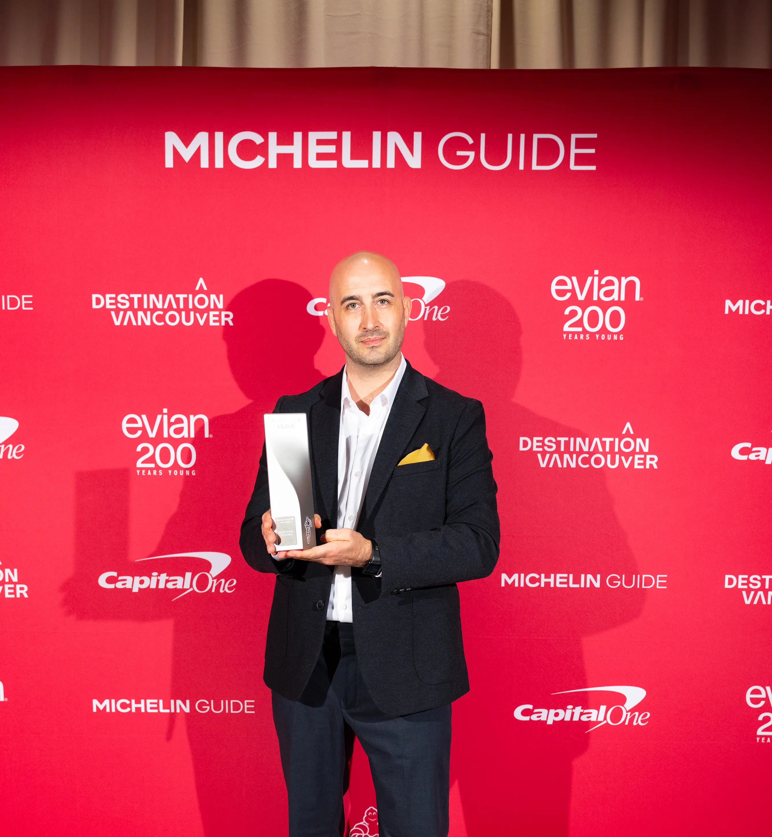 Chris Enns, the winner of the MICHELIN Exceptional Cocktails award, in a black suit holding a Michelin Guide award in front of a red backdrop with Michelin Guide and sponsor logos.