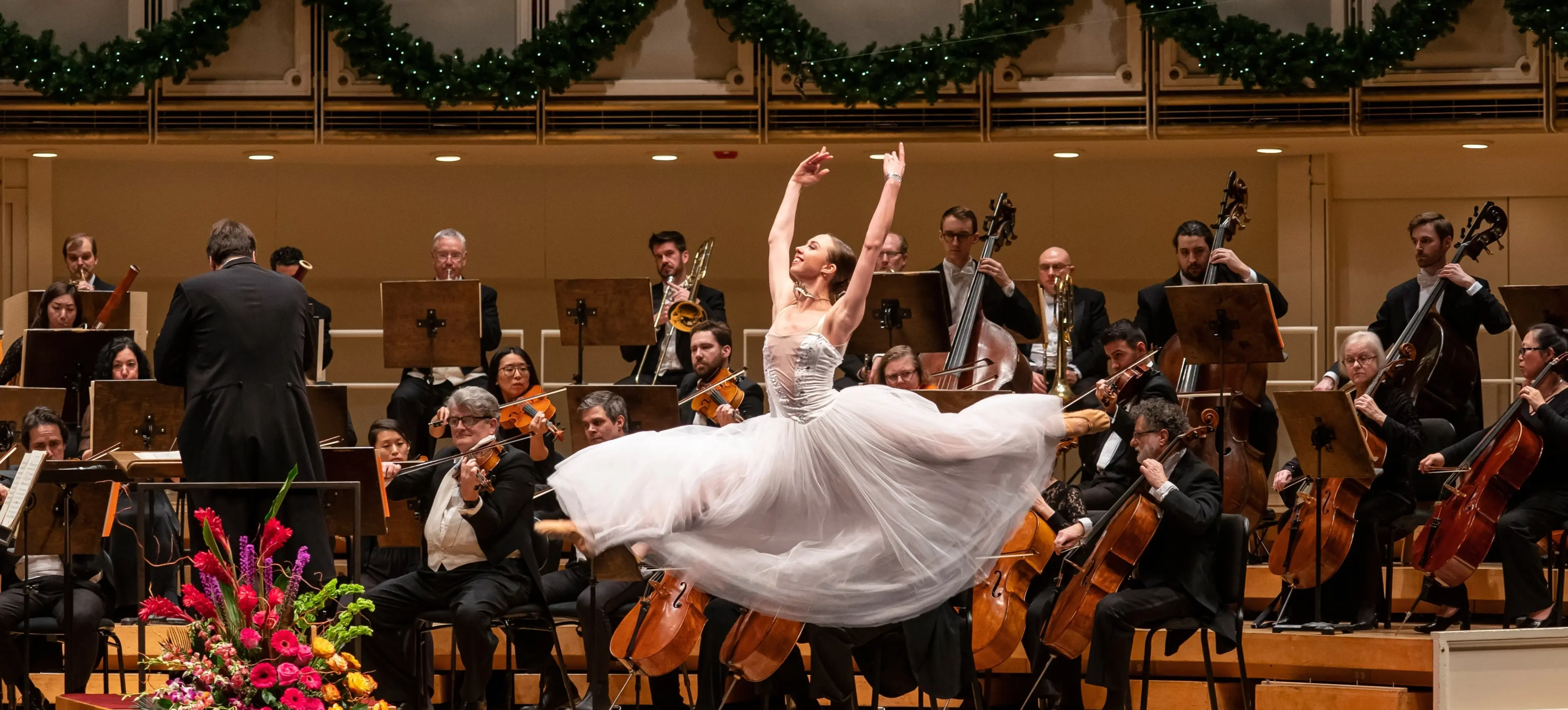 Ballet dancer in a white dress performing a grand jeté on stage with an orchestra playing behind.