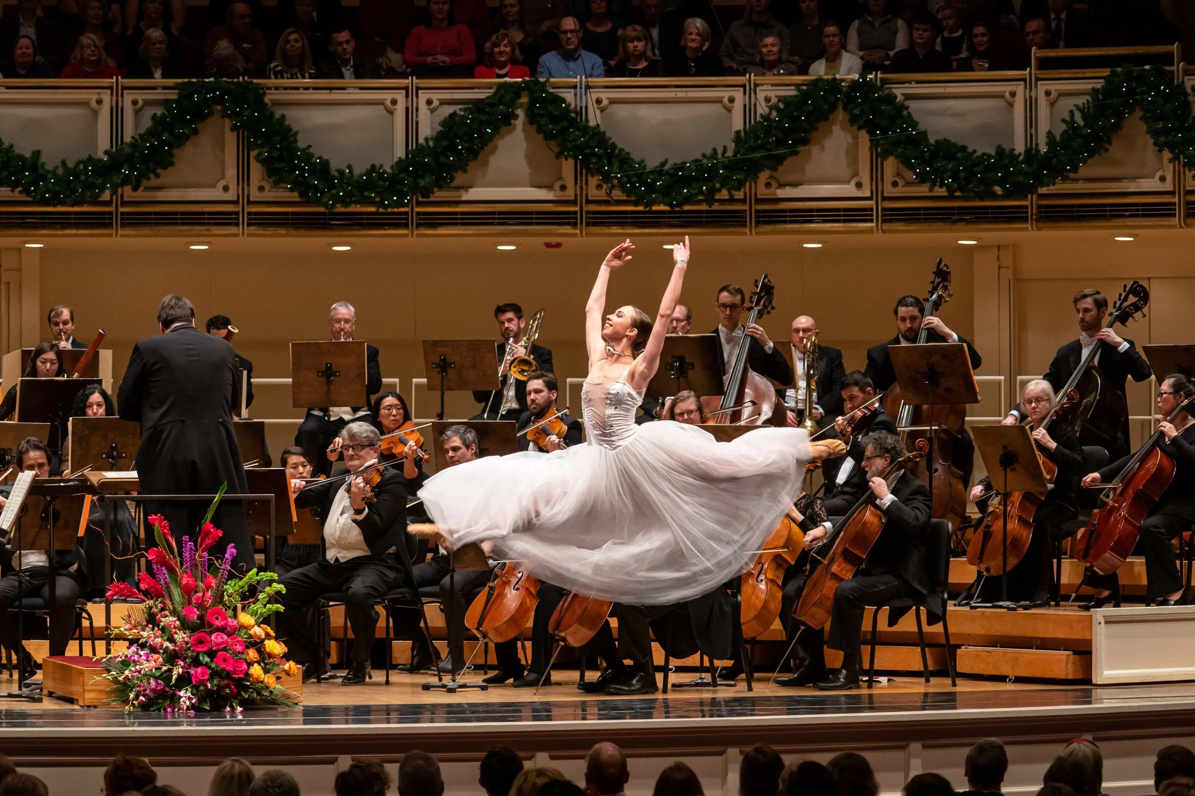 Ballet dancer in a white dress performing a grand jeté on stage with an orchestra playing behind.