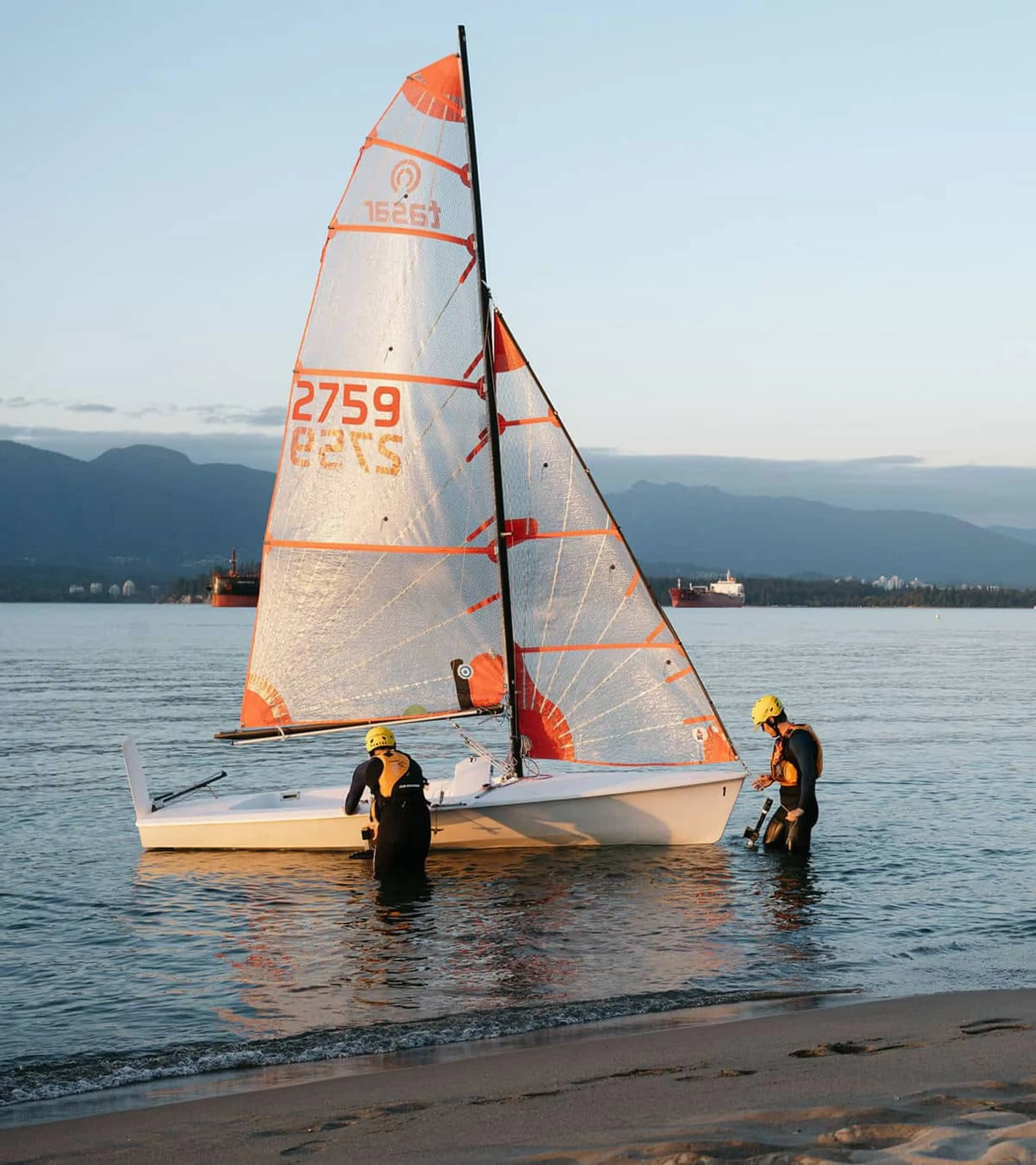 Two people in wetsuits and yellow helmets preparing a small sailboat with orange and white sails near a sandy shore at sunset.