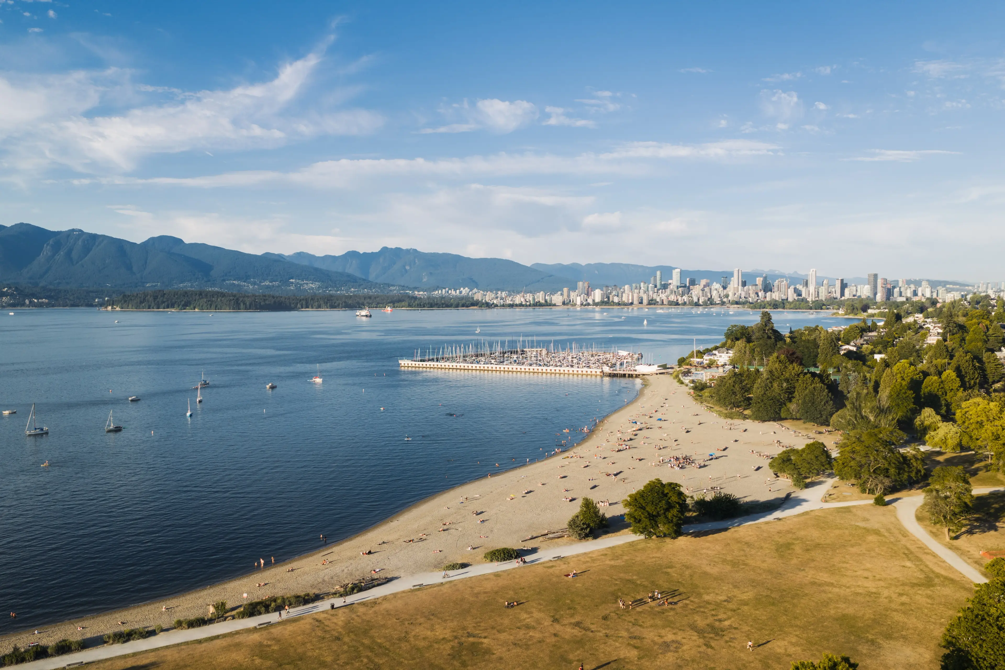 A sandy beach with scattered people, a marina with boats, a city skyline in the distance, and mountains under a partly cloudy sky.