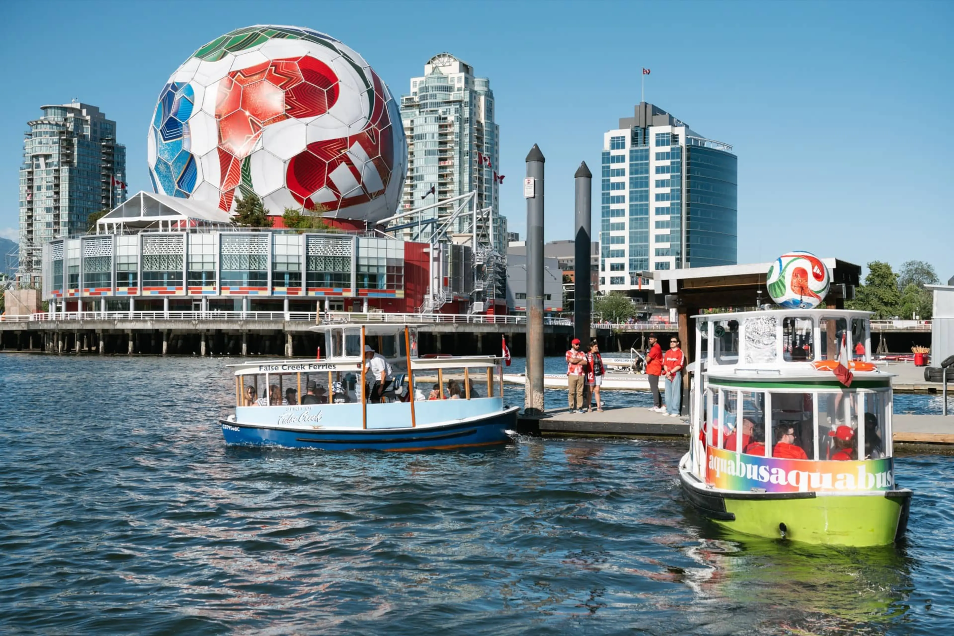 Two small passenger ferries on water near a dock with people, with a large building topped by a giant soccer ball in the background.