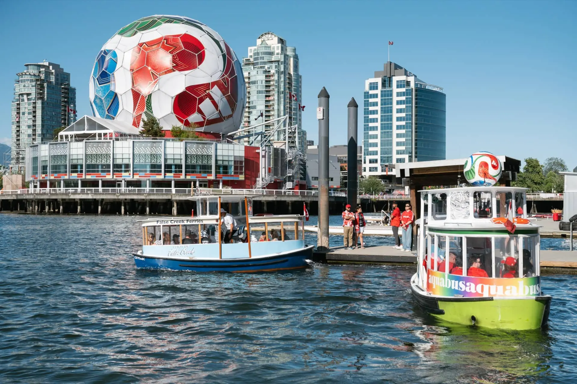 Two small passenger ferries on water near a dock with people, with a large building topped by a giant soccer ball in the background.