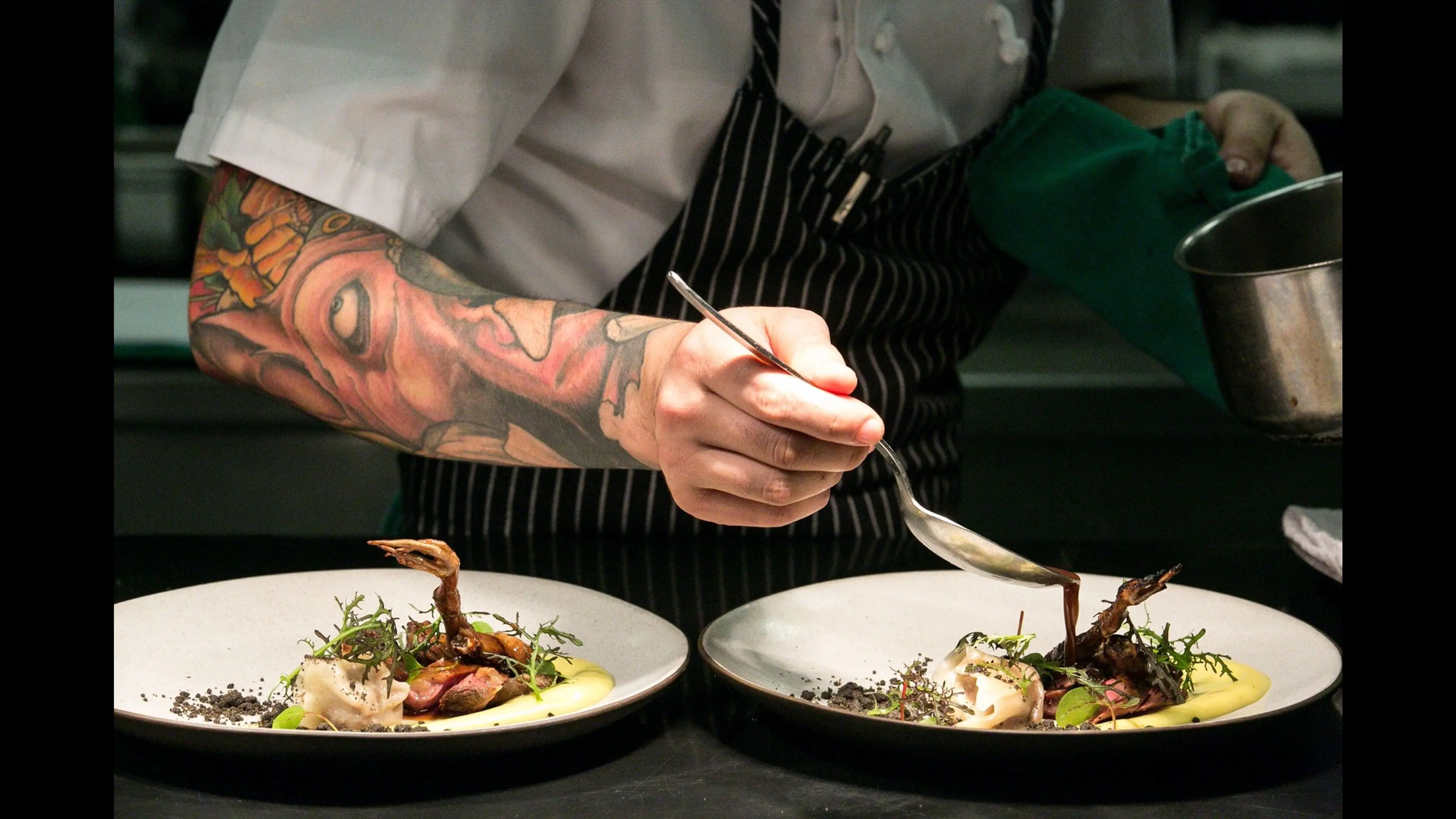A tattooed chef in a striped apron carefully drizzles sauce onto two plated gourmet dishes with greens and a small bird garnish.